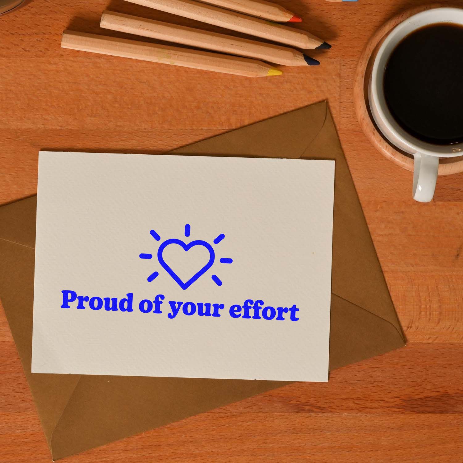 Blue heart stamp and seal on white card reading Proud of your effort, with brown envelope, pencils, and coffee.