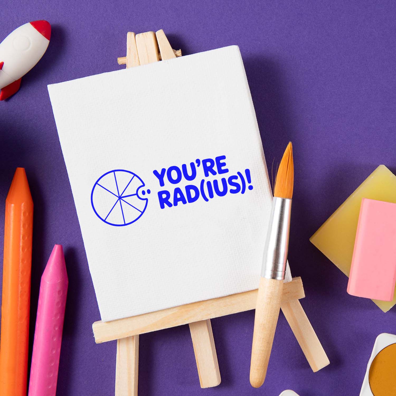 Blue custom stamp and seal on a white canvas easel, saying YOURE RAD(IUS)! on a purple background with art supplies.