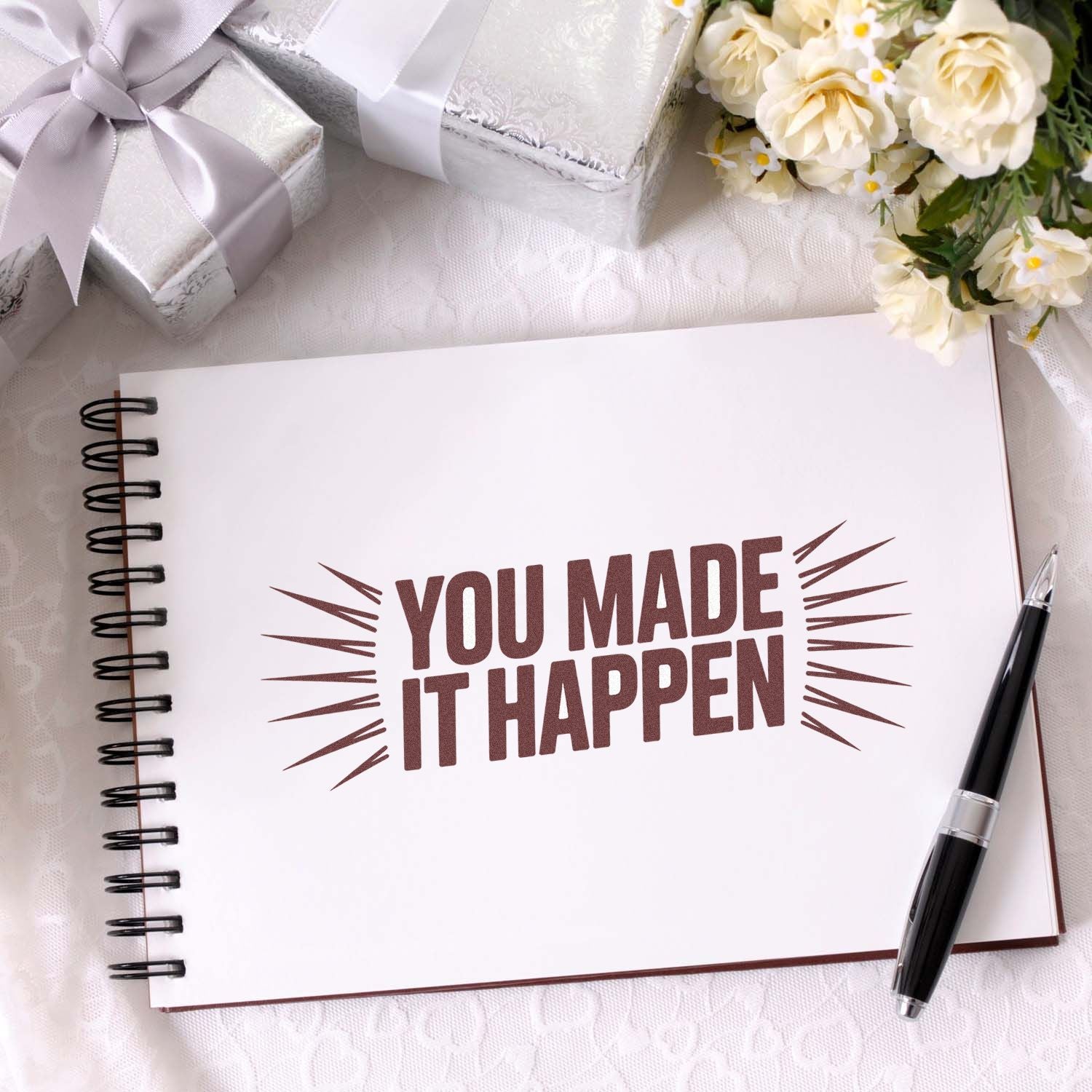 Brown "You Made It Happen" stamp on white spiral notebook, with silver gift boxes, cream flowers, and black pen.