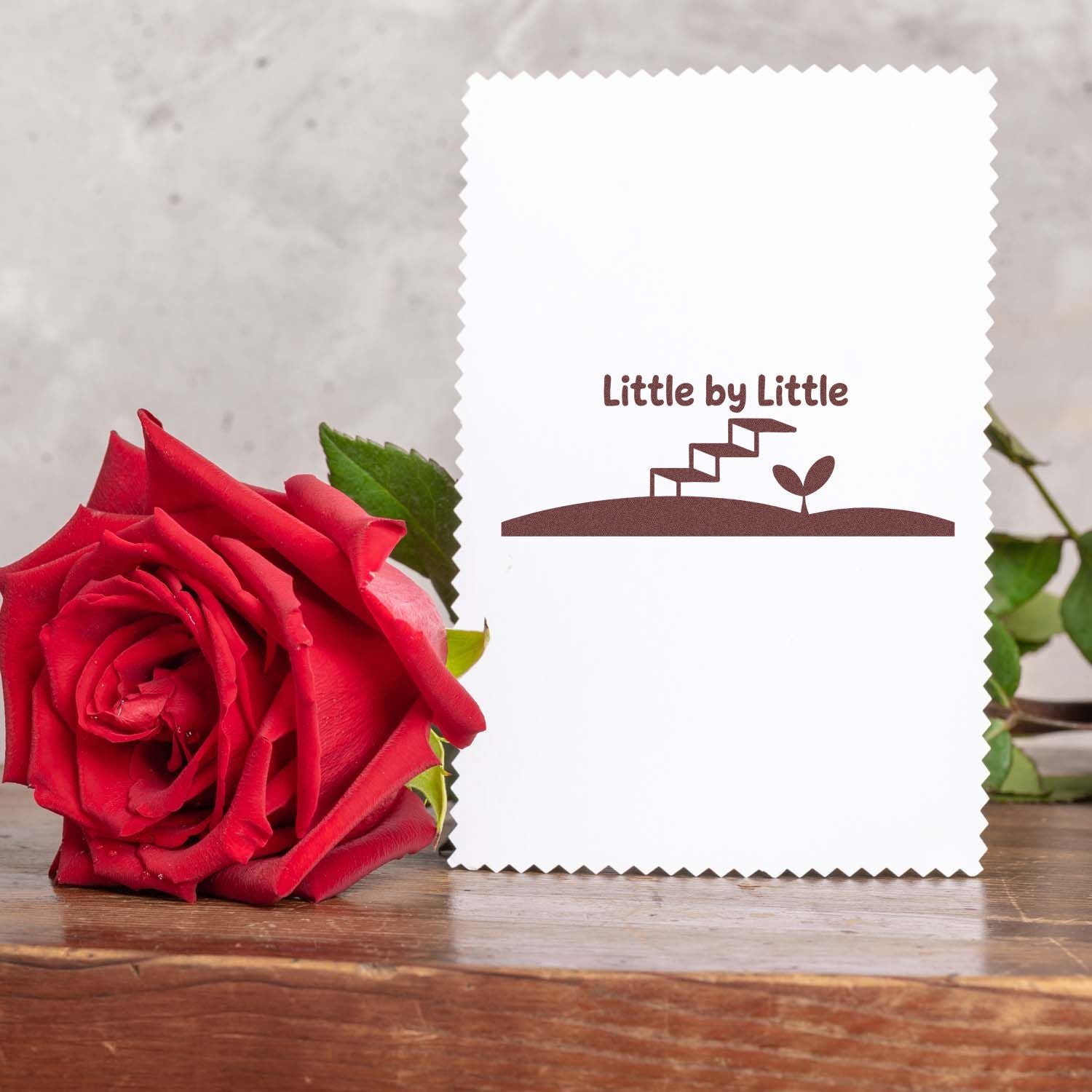 White scalloped stamp card with brown Little by Little seal, staircase sprout logo, beside a red rose.