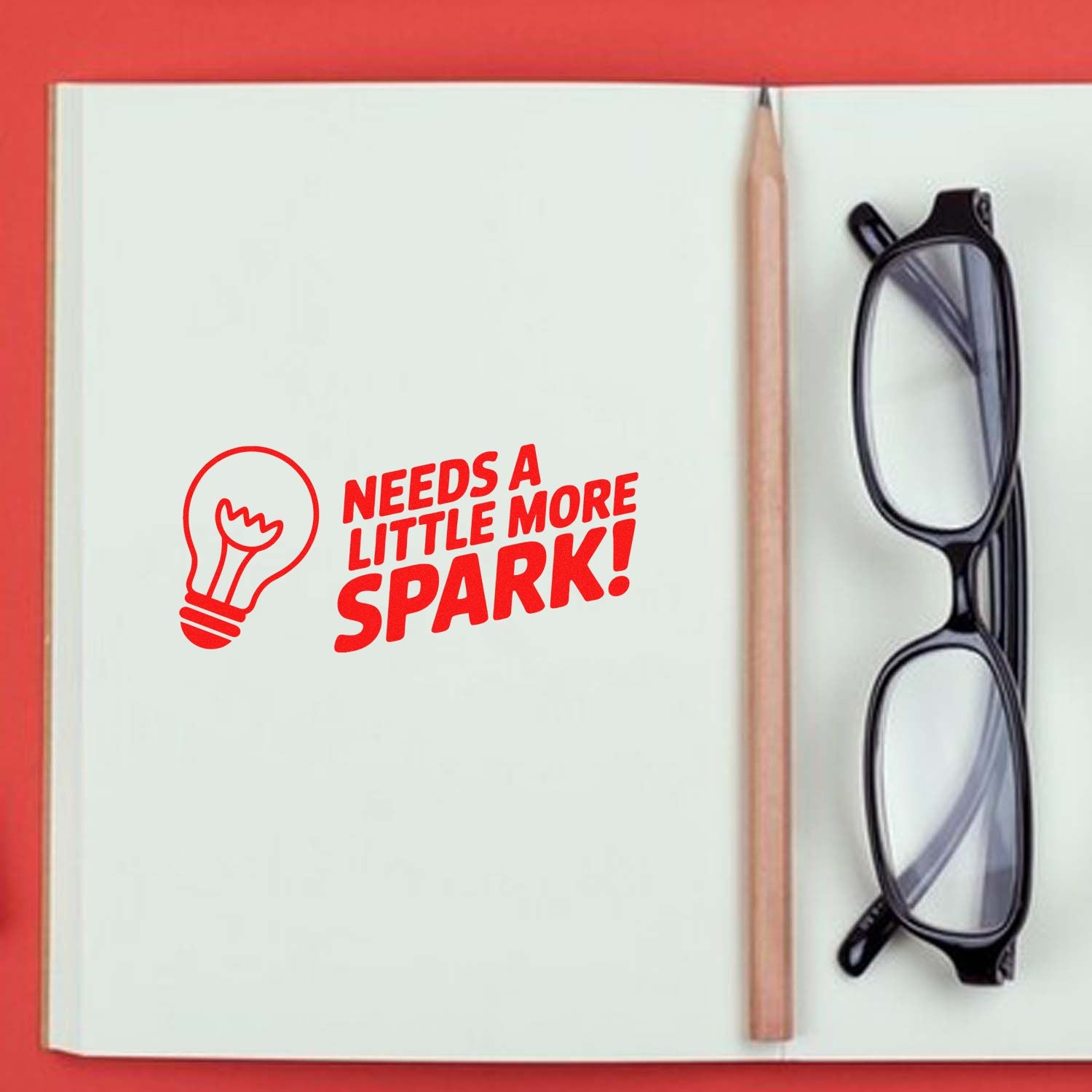 Red custom stamp with lightbulb and Needs a Little More Spark! on open white notebook, beside pencil and black glasses.