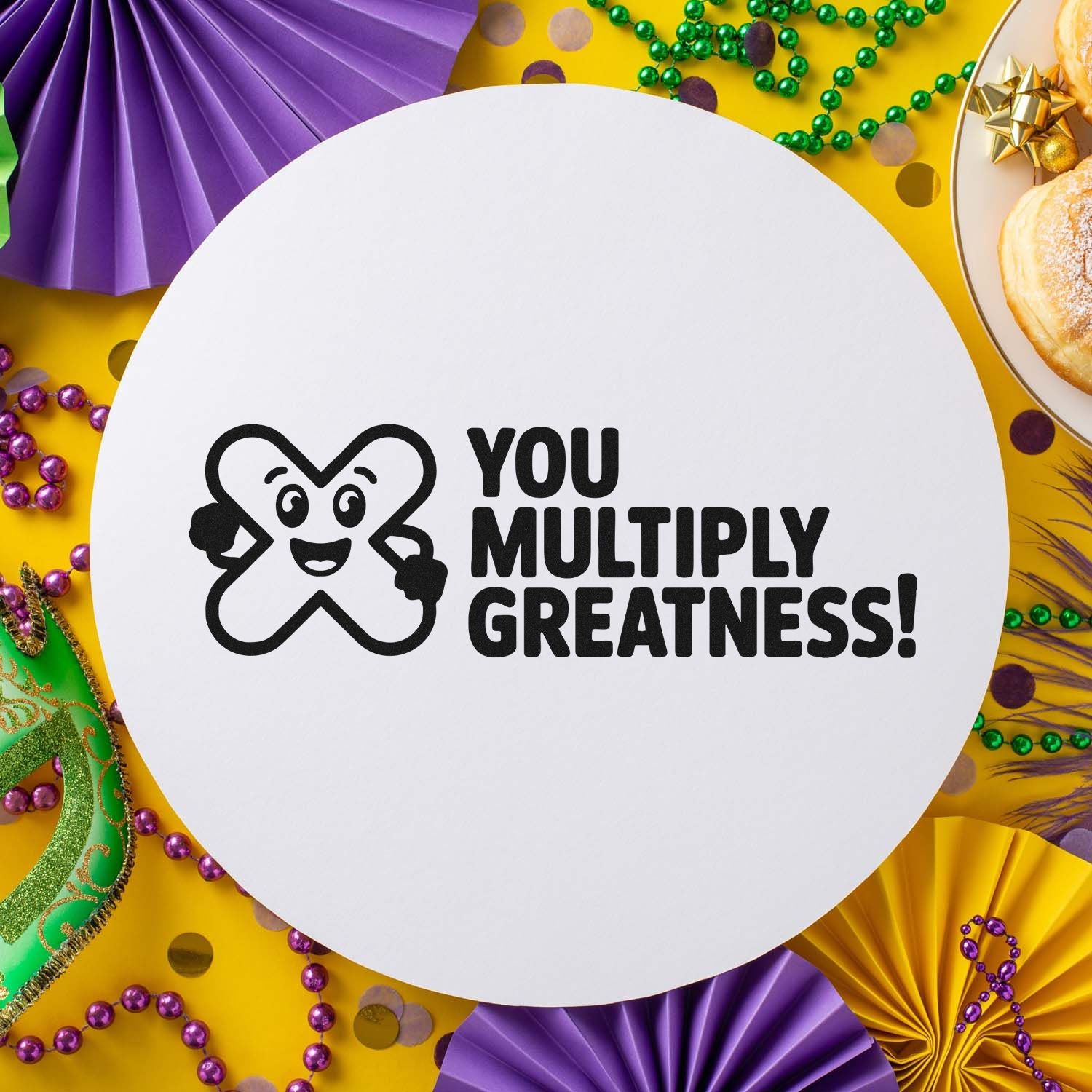 White round stamp seal with black You Multiply Greatness! text on a yellow Mardi Gras background with purple, green and gold décor.