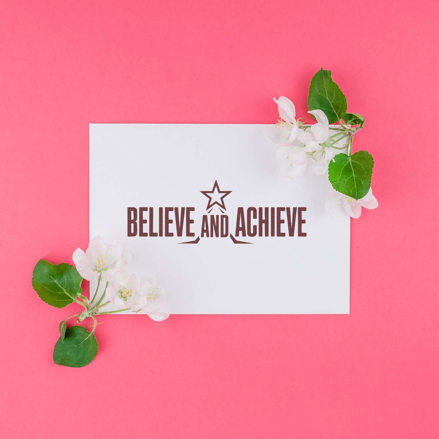 White stamp and seal card with brown Believe and Achieve text, star logo, on pink background with white blossoms and green leaves.