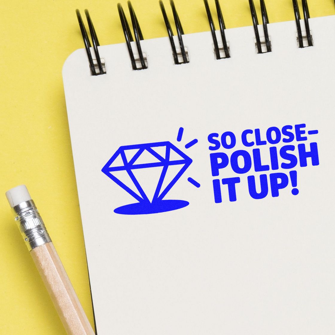 Blue stamp and seal design with diamond and So Close- Polish It Up! on a white spiral notebook, beside a pencil on yellow background.