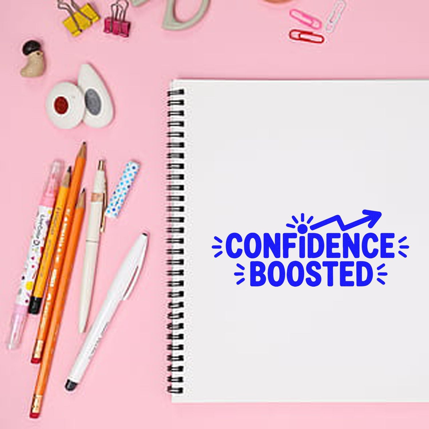 Blue CONFIDENCE BOOSTED stamp on a white spiral notebook, surrounded by pink stationery on a pastel pink background.