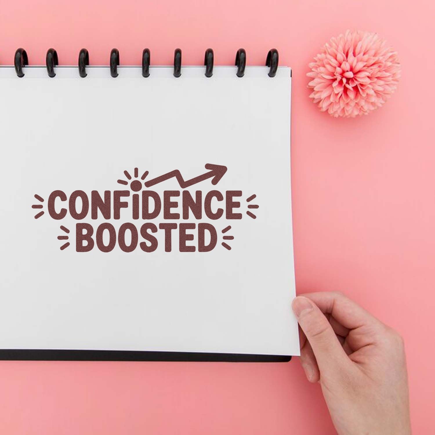 Pink notebook with a brown Confidence Boosted stamp, arrow graphic, and flower accent on a pastel pink background.