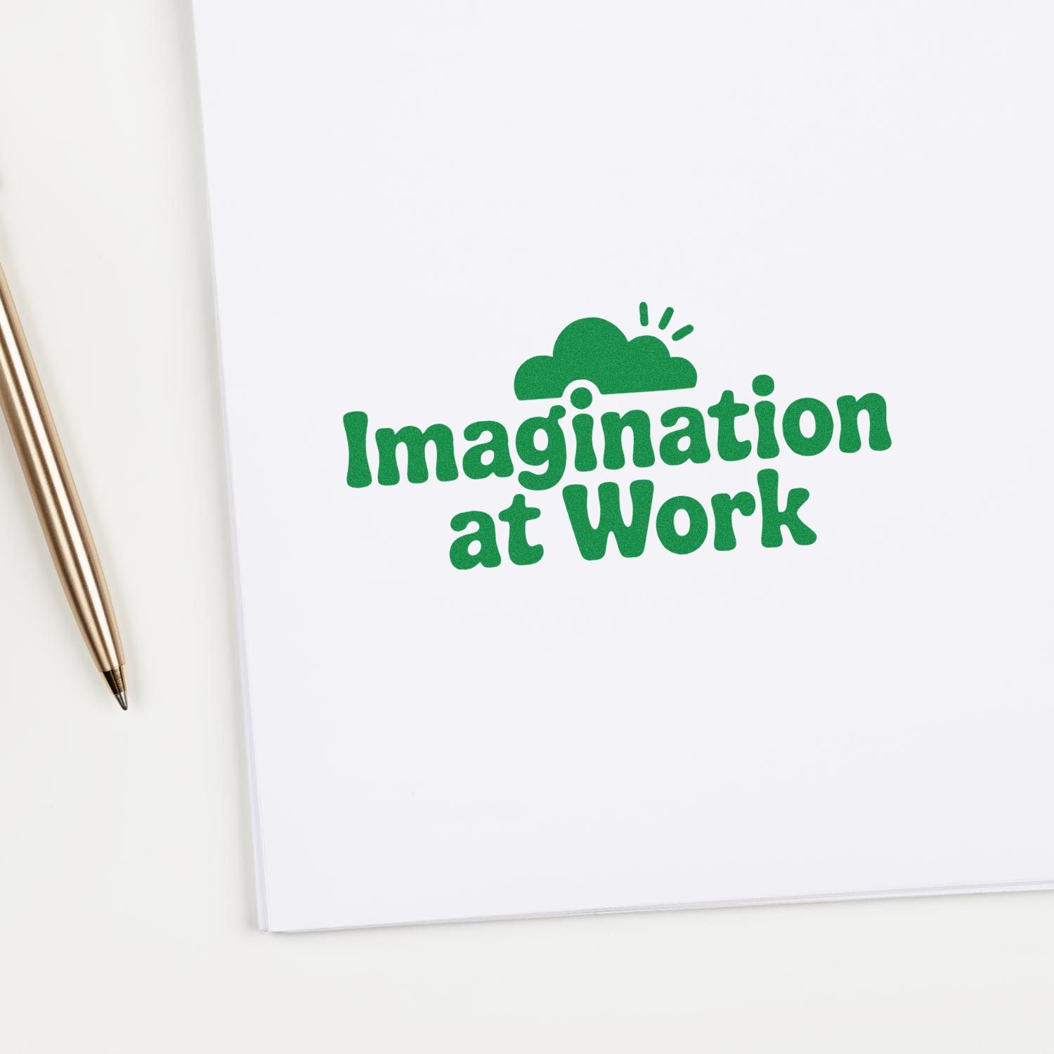 Green stamp seal reading Imagination at Work on white paper, with a gold pen beside it.