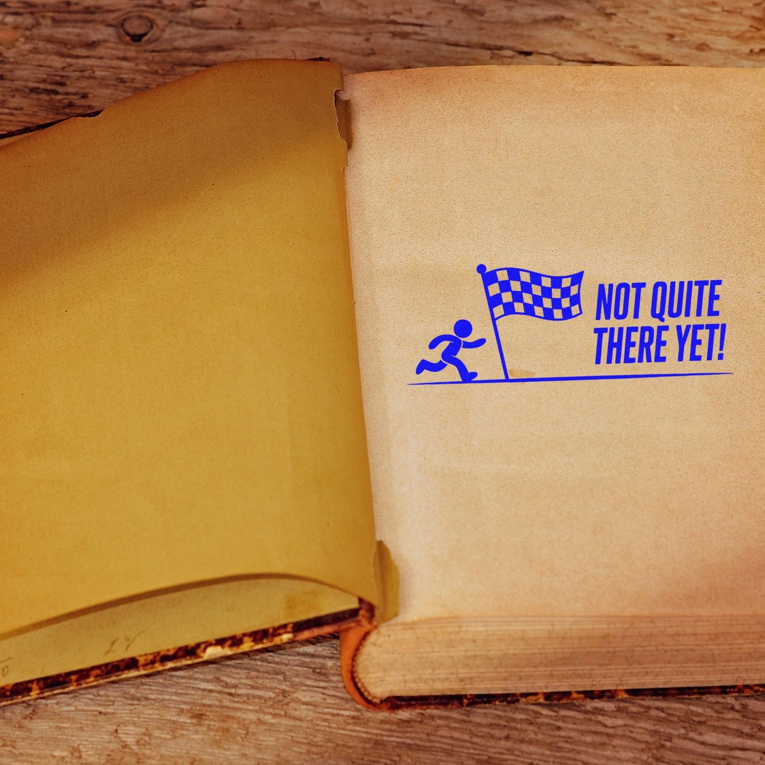 Blue stamp seal with runner and checkered flag saying Not Quite There Yet! on an open tan book.
