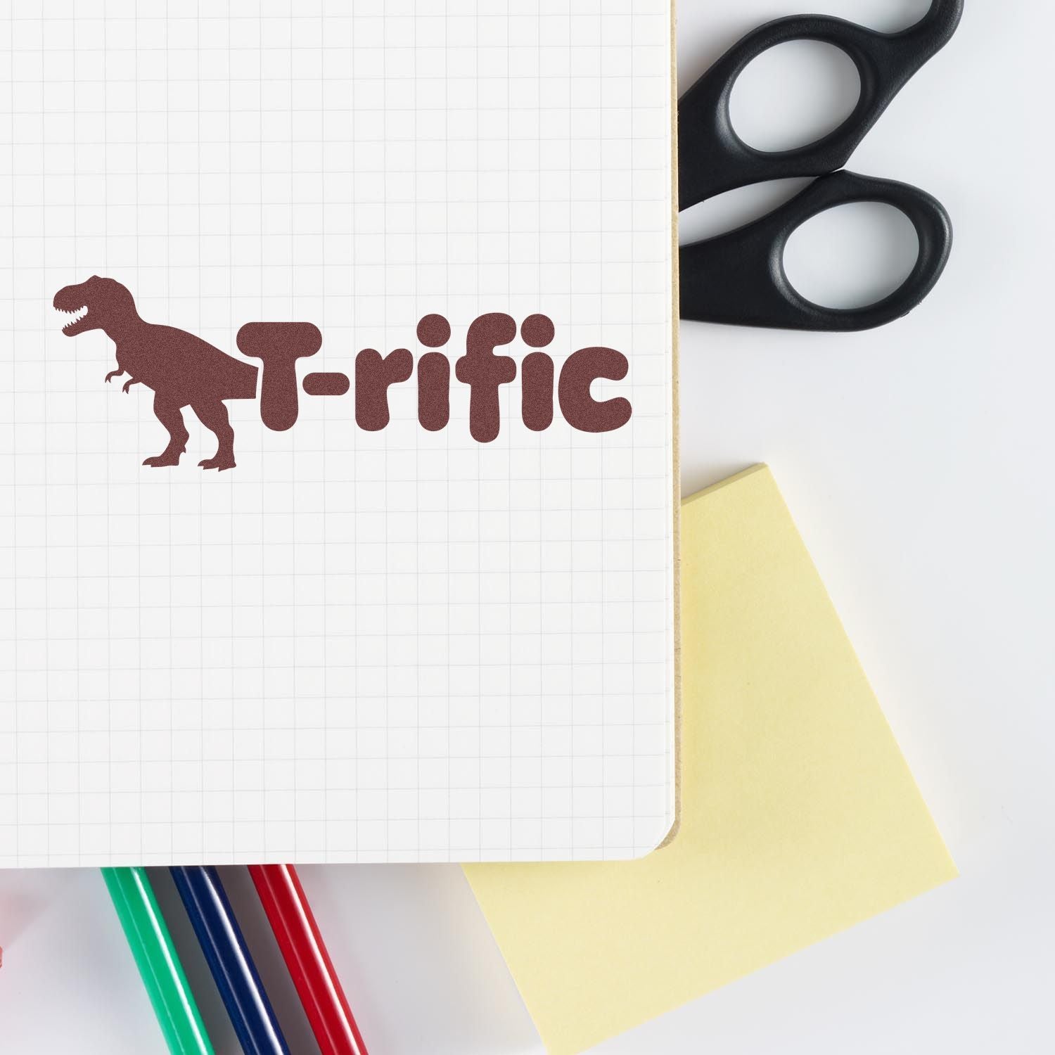 Brown T-rific dinosaur custom stamp/seal on grid notebook, with black scissors, yellow note, and colored pens.