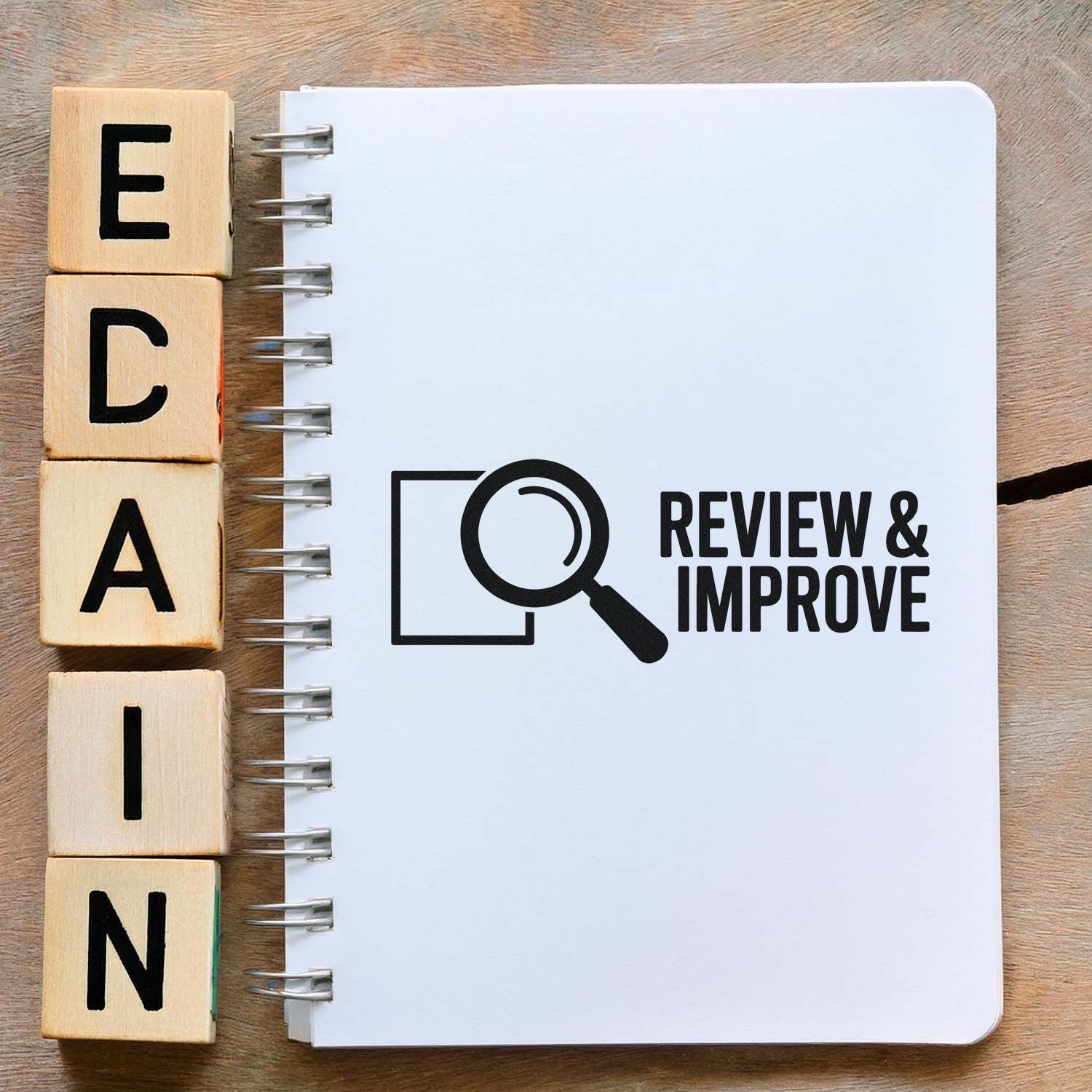Wooden stamp blocks spelling ECAIN beside a white spiral notebook with a black Review & Improve seal and magnifying glass.