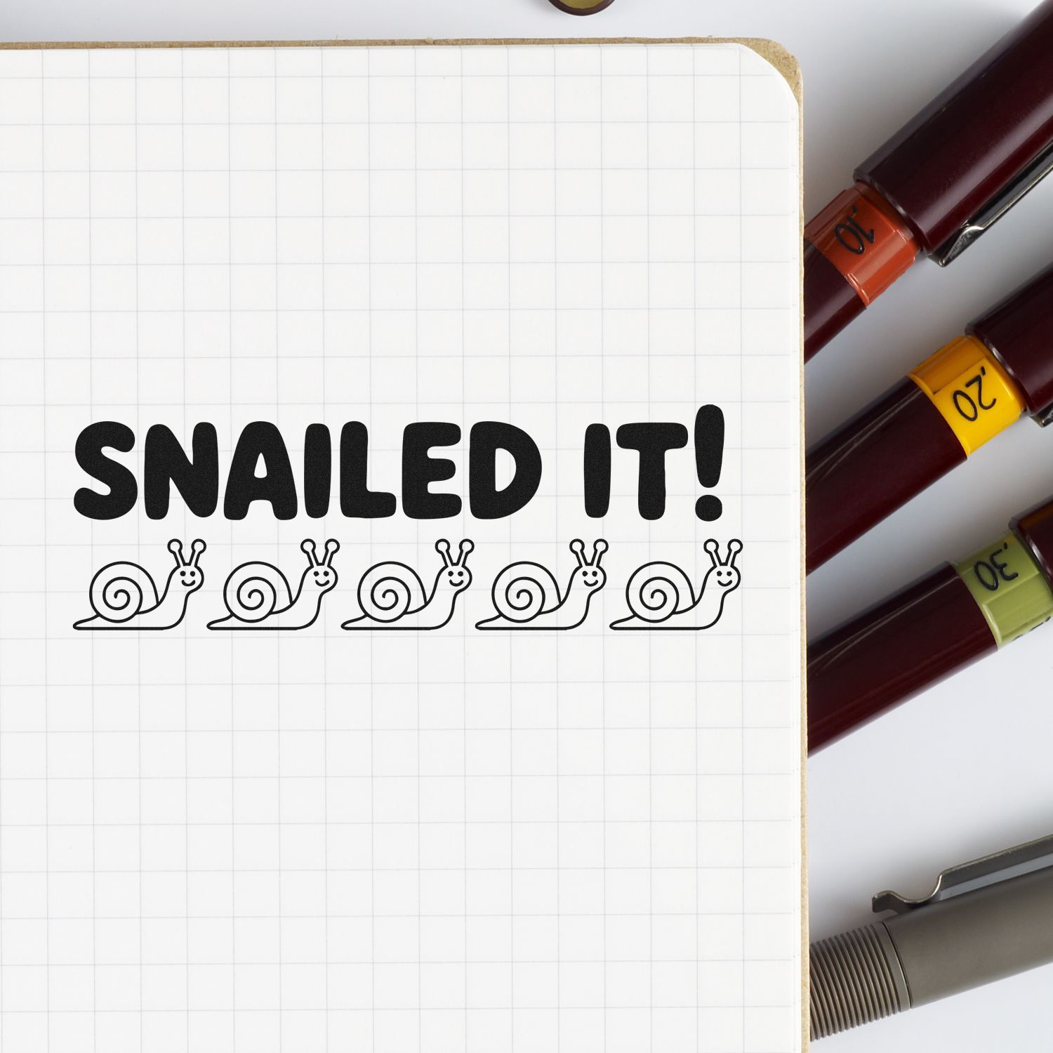 Black SNAILED IT! stamp on grid paper with snail icons, beside burgundy stamp and seal markers in orange, yellow, and green.