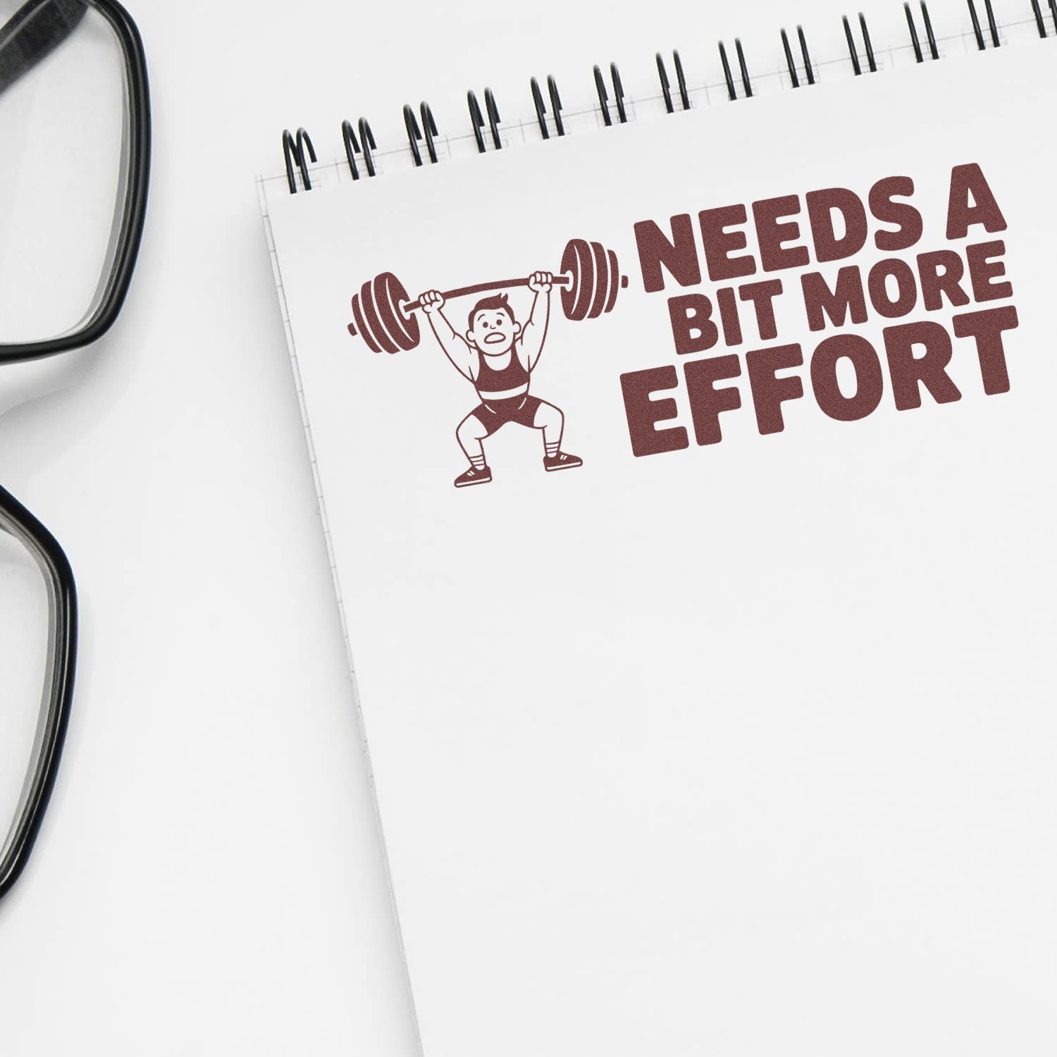 Maroon rubber stamp design on white notebook with weightlifter and NEEDS A BIT MORE EFFORT text, beside black eyeglasses.