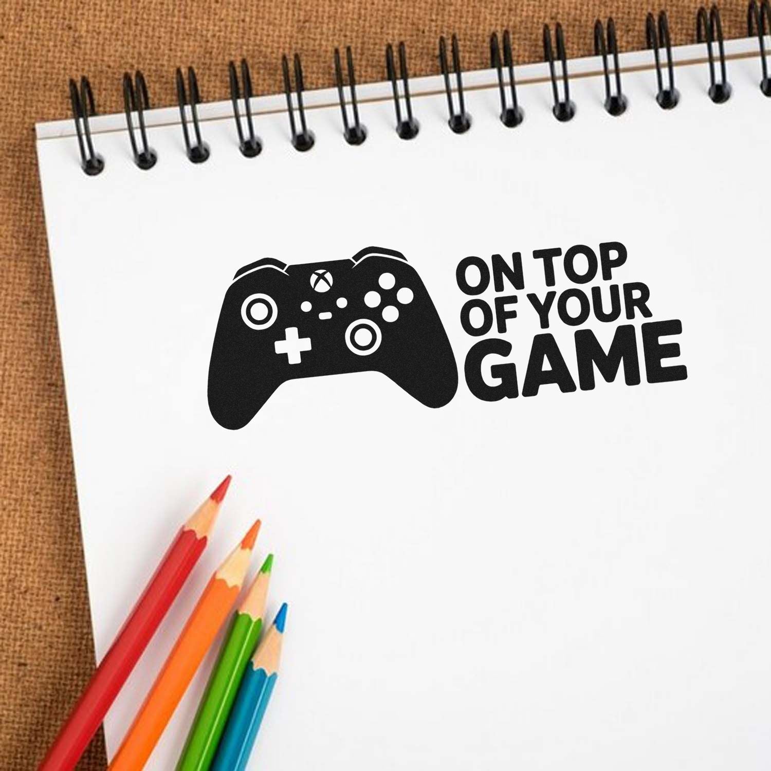Black game controller stamp and seal on white spiral notebook, with On Top of Your Game text and colored pencils.