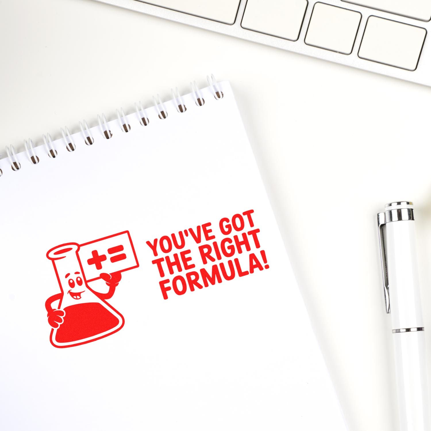 Red self-inking stamp/seal on white notebook says Youve got the right formula! with beaker graphic; white pen and keyboard.