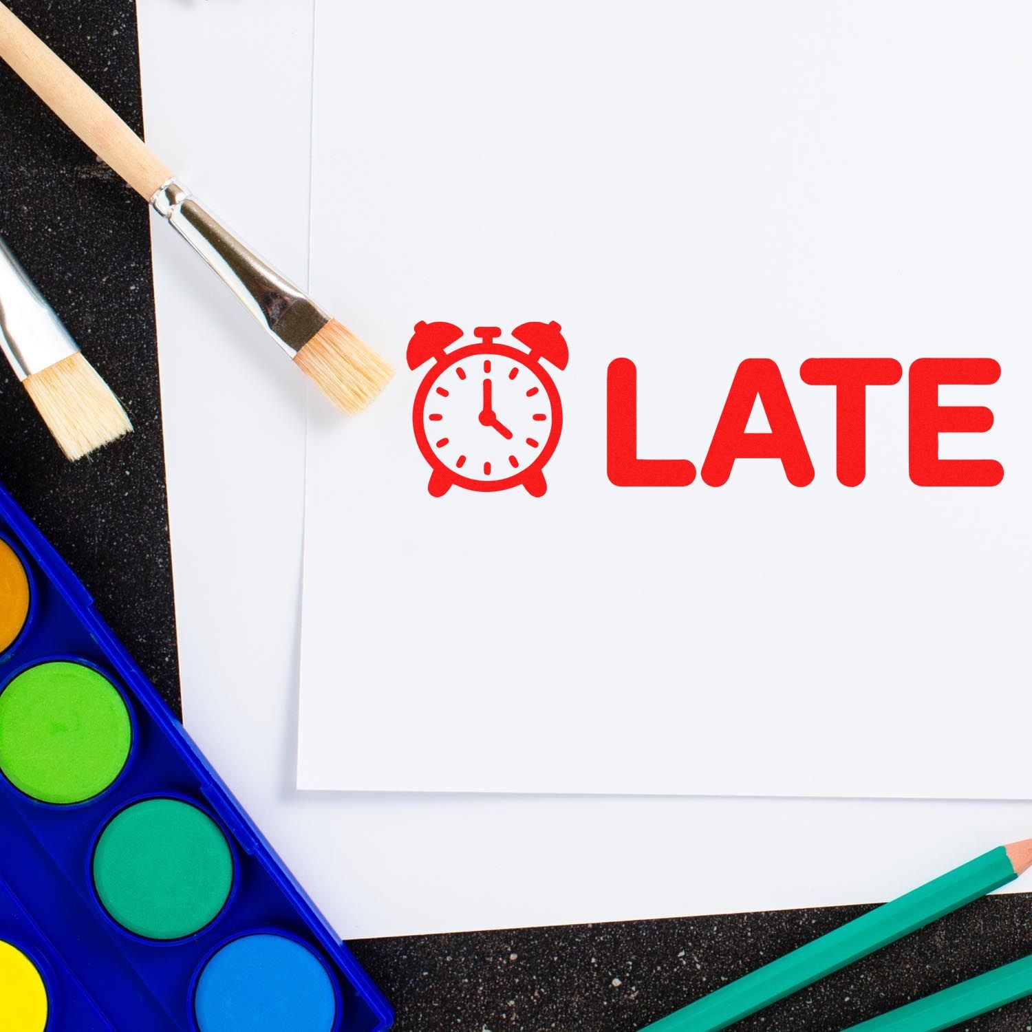 Red LATE stamp with alarm clock icon on white paper, beside paint brushes, pencils, and a blue watercolor palette.