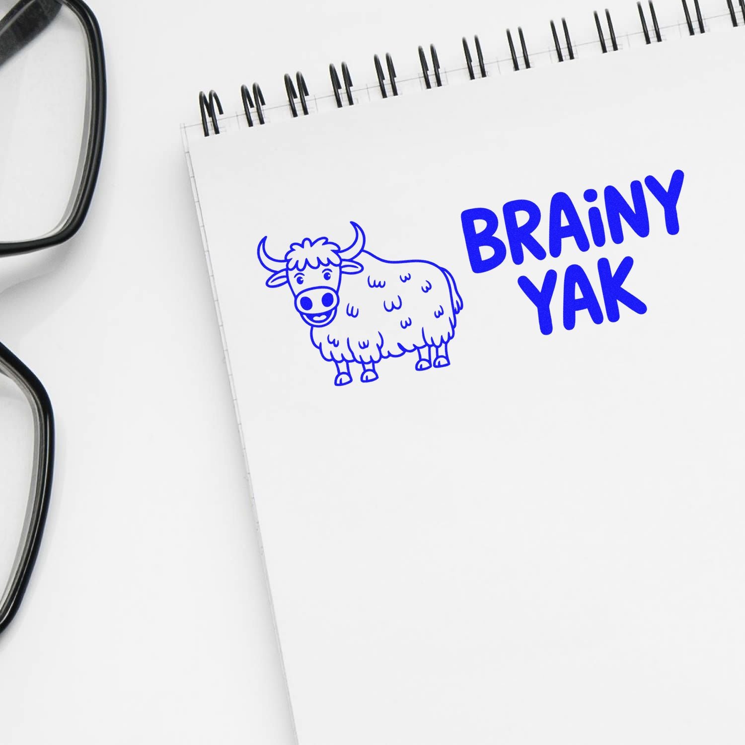 Blue BRAINY YAK stamp with cartoon cow on white spiral notebook, beside black eyeglasses.