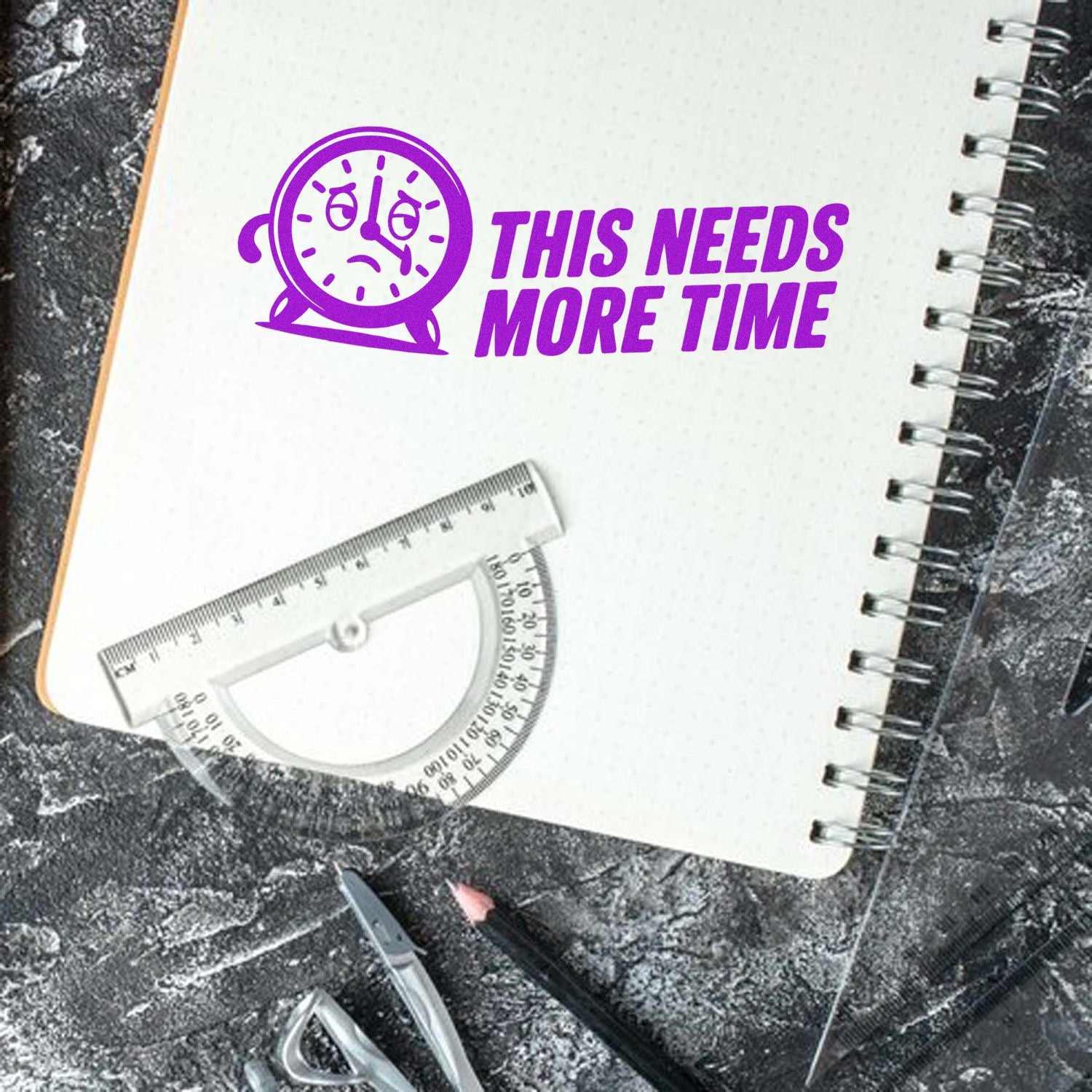 Purple self-inking stamp seal with sad clock and THIS NEEDS MORE TIME on a white spiral notebook