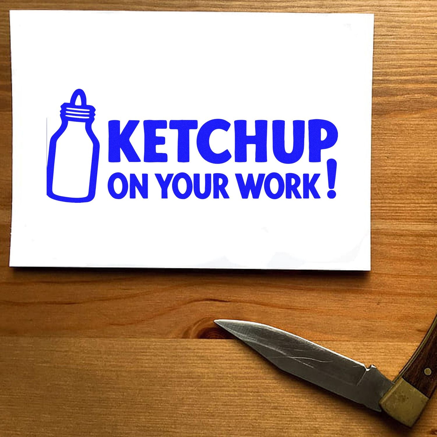 Blue ketchup bottle rubber stamp reading KETCHUP ON YOUR WORK! on white paper, with a knife on a wood table.
