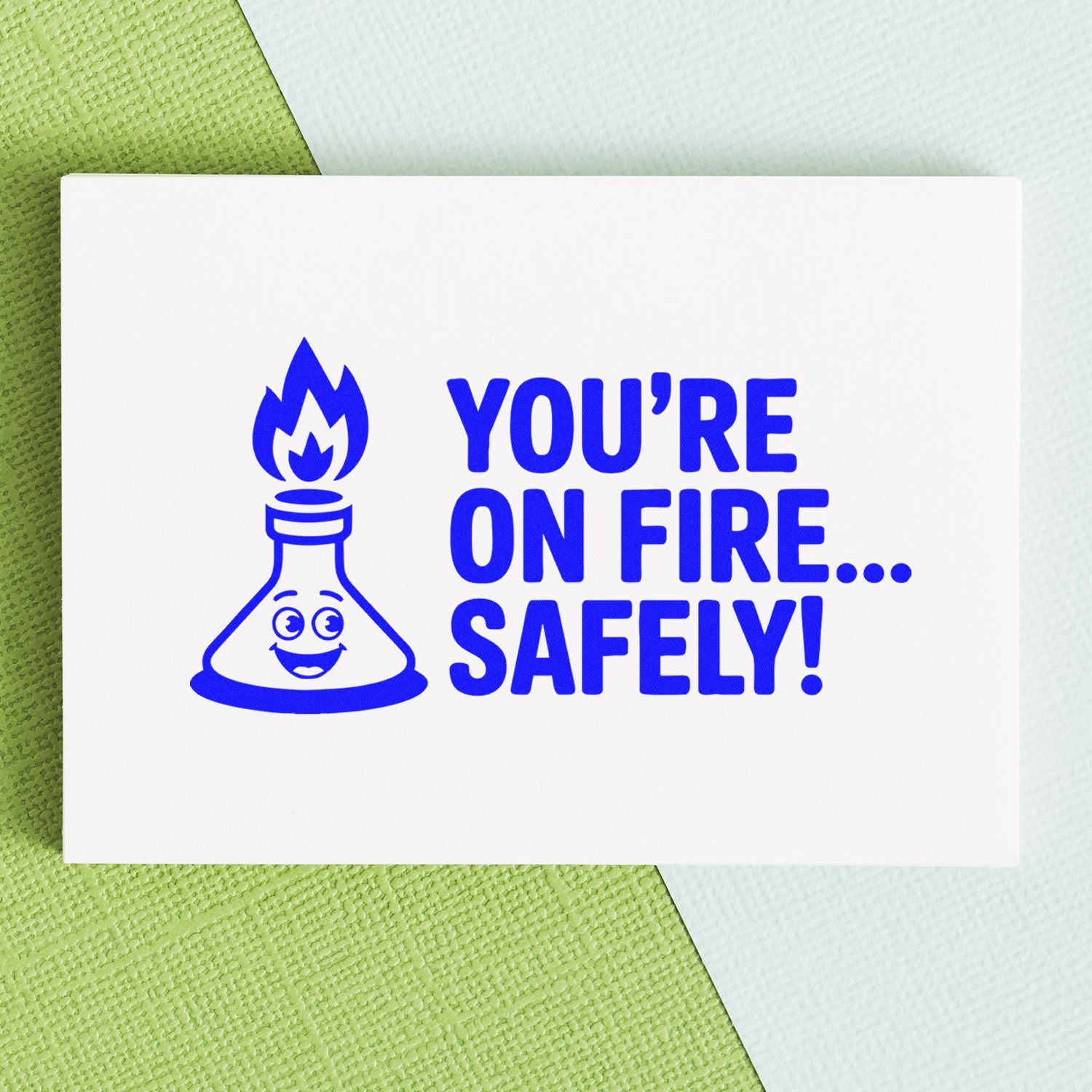 Blue rubber stamp seal on white card with smiling flask and flame, reading Youre on fire... safely! on green and white background.