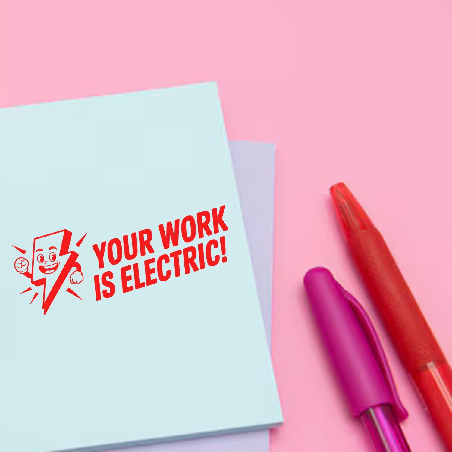 Red electric-themed stamp and seal reading YOUR WORK IS ELECTRIC! on pale blue paper, with red and purple pens on pink.