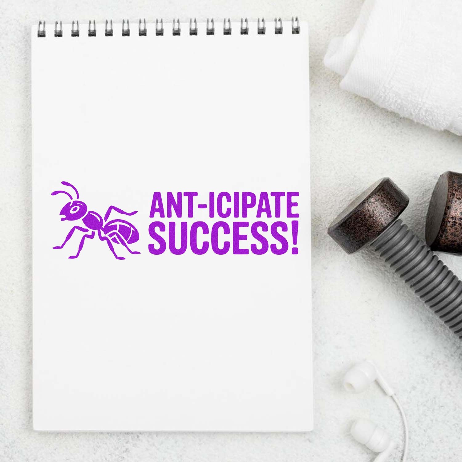 Purple ant-themed stamp on white spiral notebook reading ANT-ICIPATE SUCCESS, with black stamp and seal tools nearby.