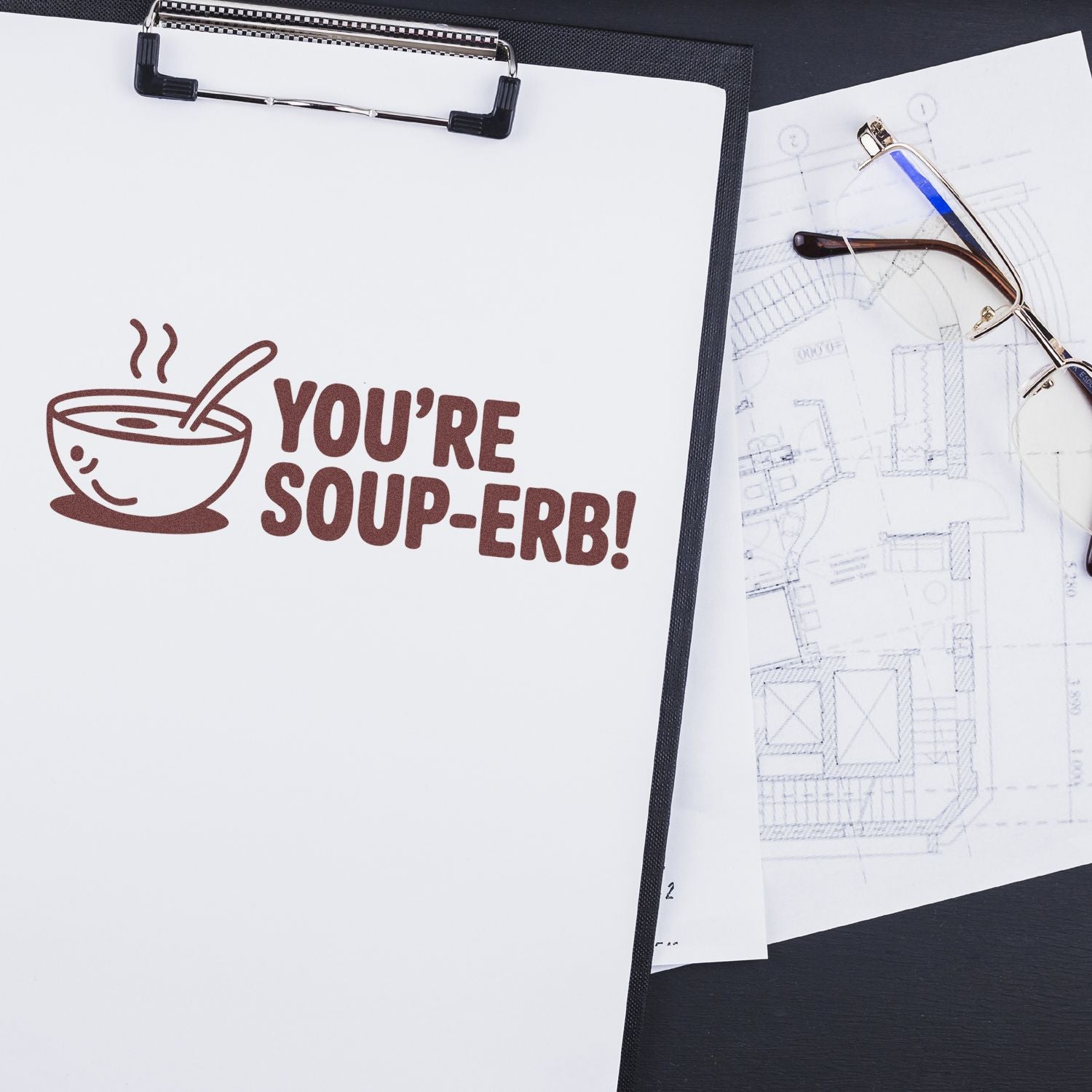 Custom brown stamp reading YOURE SOUP-ERB! on white clipboard, beside eyeglasses and architectural plans.