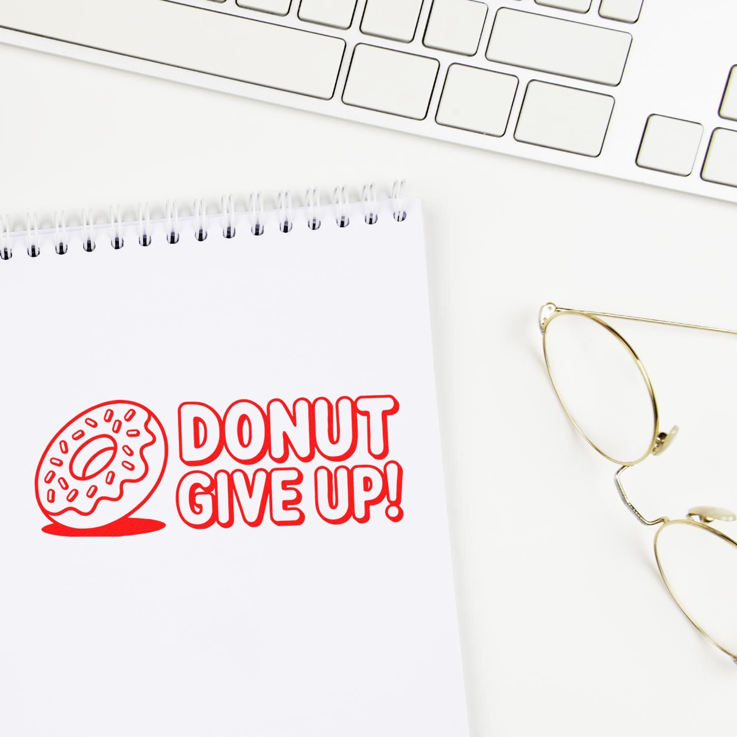 Red donut stamp seal reading DONUT GIVE UP! on white notepad beside gold eyeglasses and keyboard.