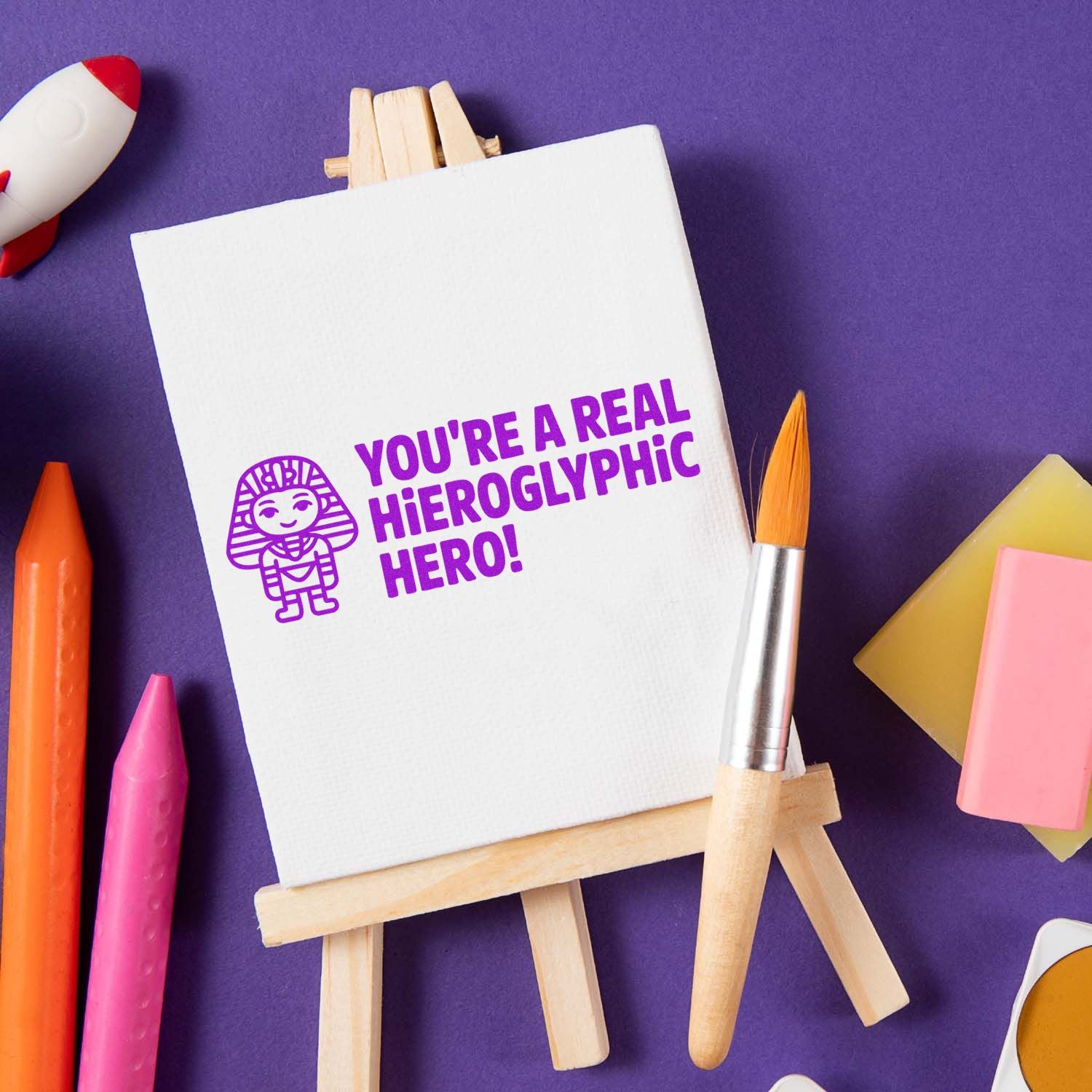 Purple stamp on white canvas easel reading Youre a real hieroglyphic hero! with hieroglyph icon, surrounded by art supplies.