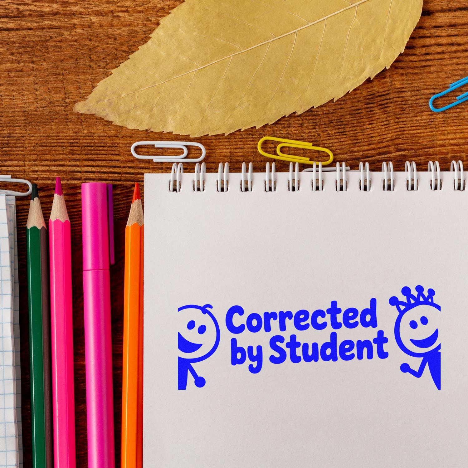 Blue Corrected by Student stamp/seal on spiral notebook with colorful pencils, paper clips, and leaf on wooden desk.