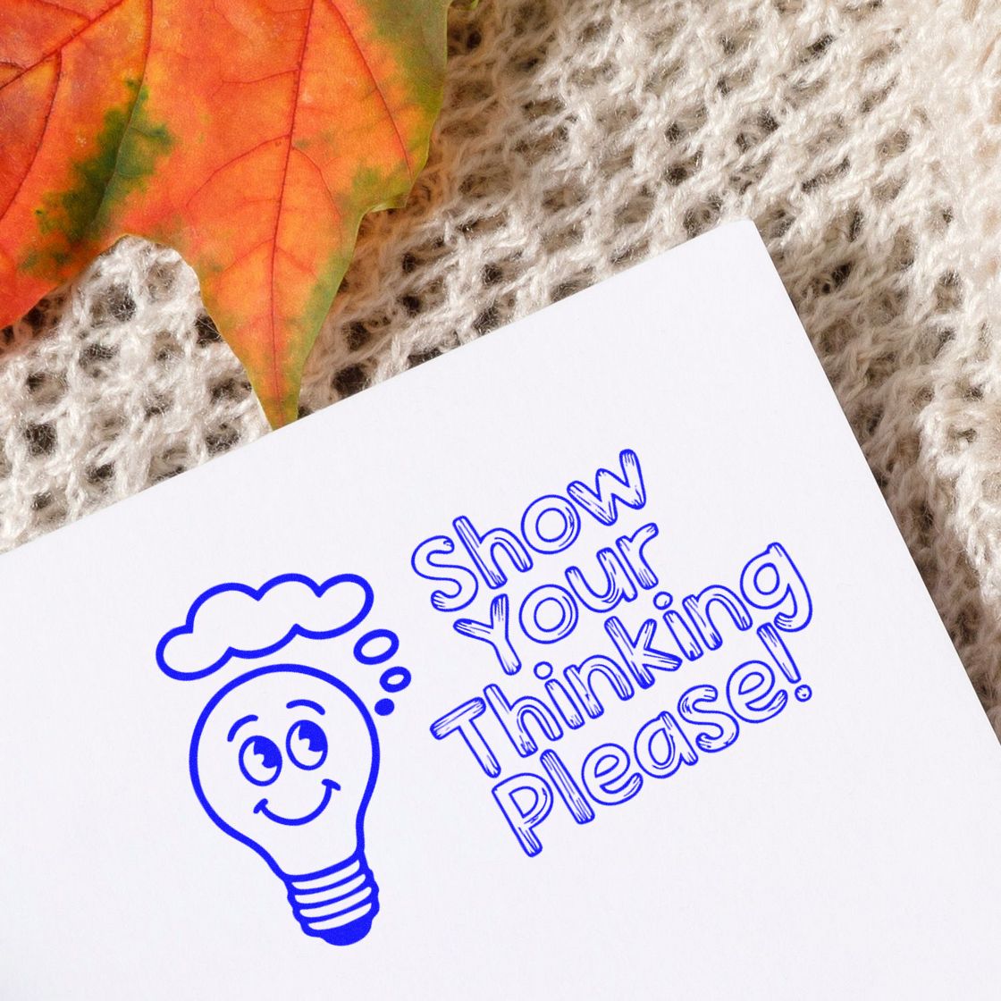 Blue custom stamp seal with smiling lightbulb and Show Your Thinking Please! on white paper, beside an orange autumn leaf.