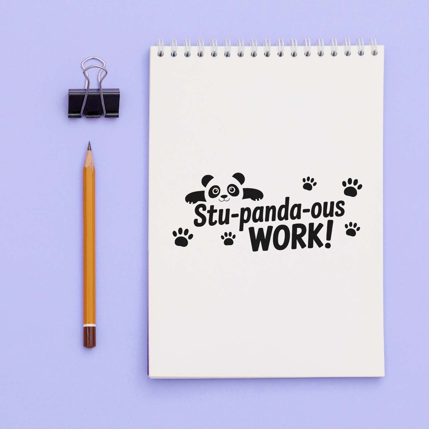 Black panda work stamp on white spiral notebook with paw prints, beside pencil and binder clip on lavender background.