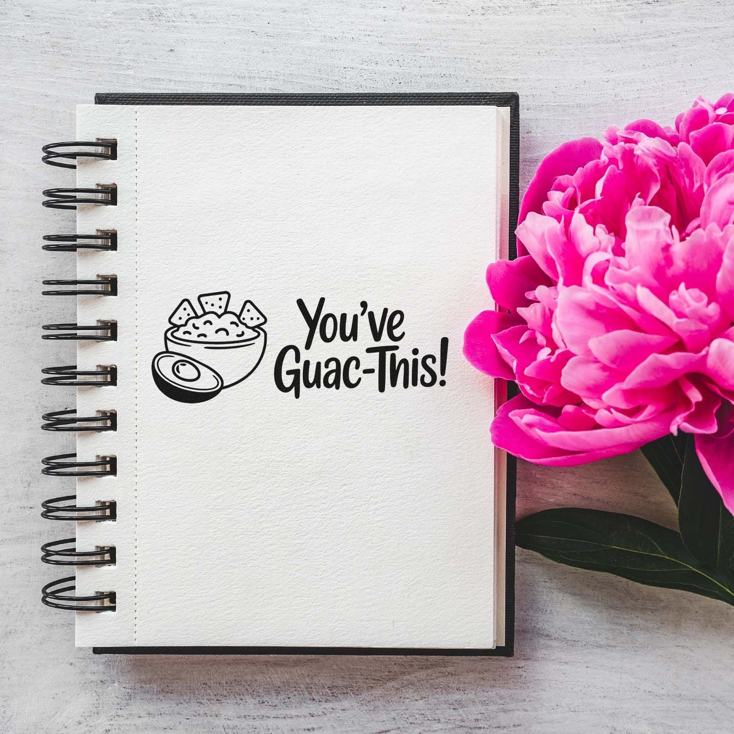 Black avocado stamp and seal reading Youve Guac-This! on white spiral notebook, with bright pink flowers.