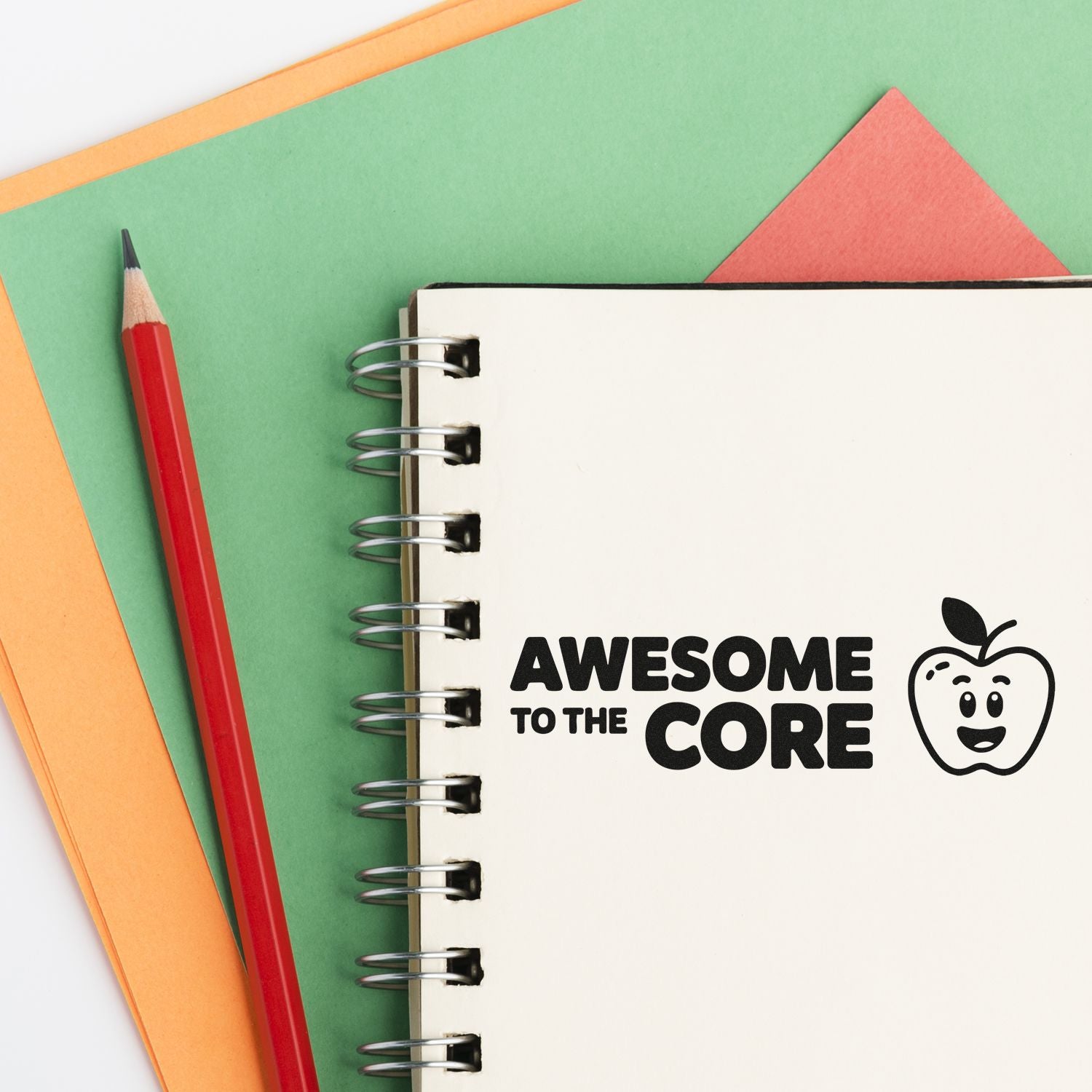 Custom stamp and seal design reading Awesome to the Core with apple icon on cream spiral notebook, red pencil, green and orange paper.