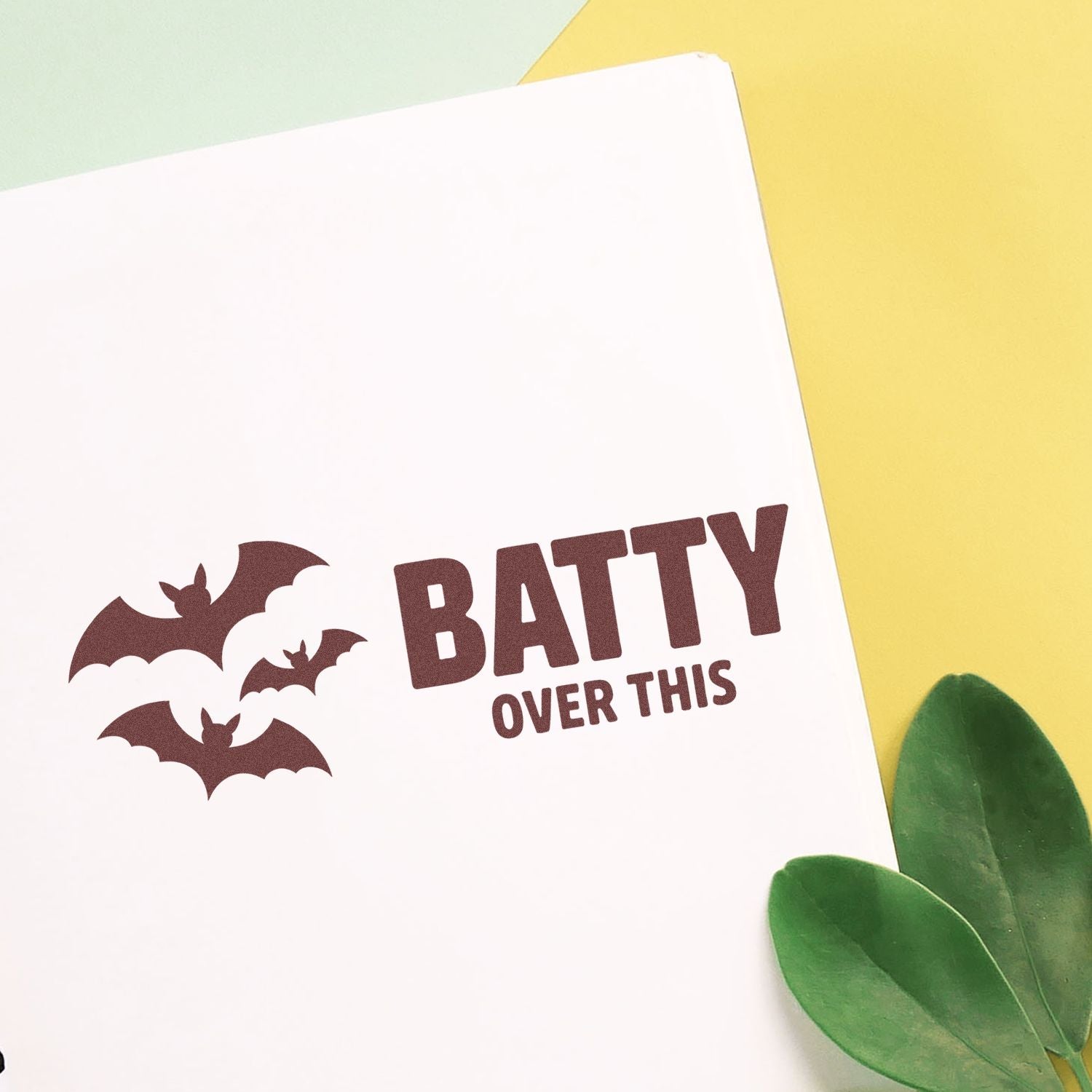 Bat-themed brown stamp and seal reading BATTY OVER THIS on a white card, with pale yellow background and green leaves.