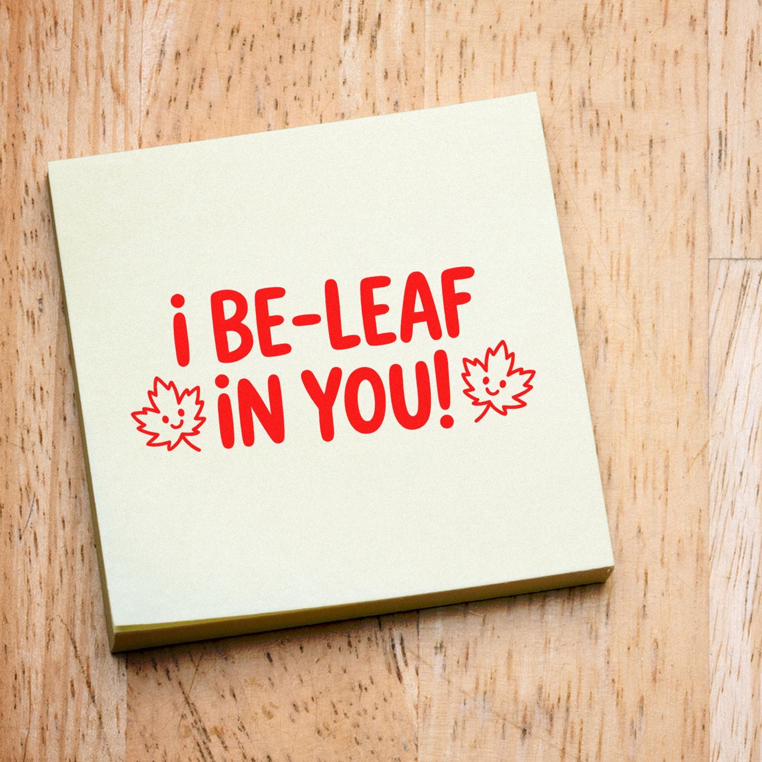 Cream square stamp seal with red I BE-LEAF IN YOU! text and smiling leaf icons on a light wood background.