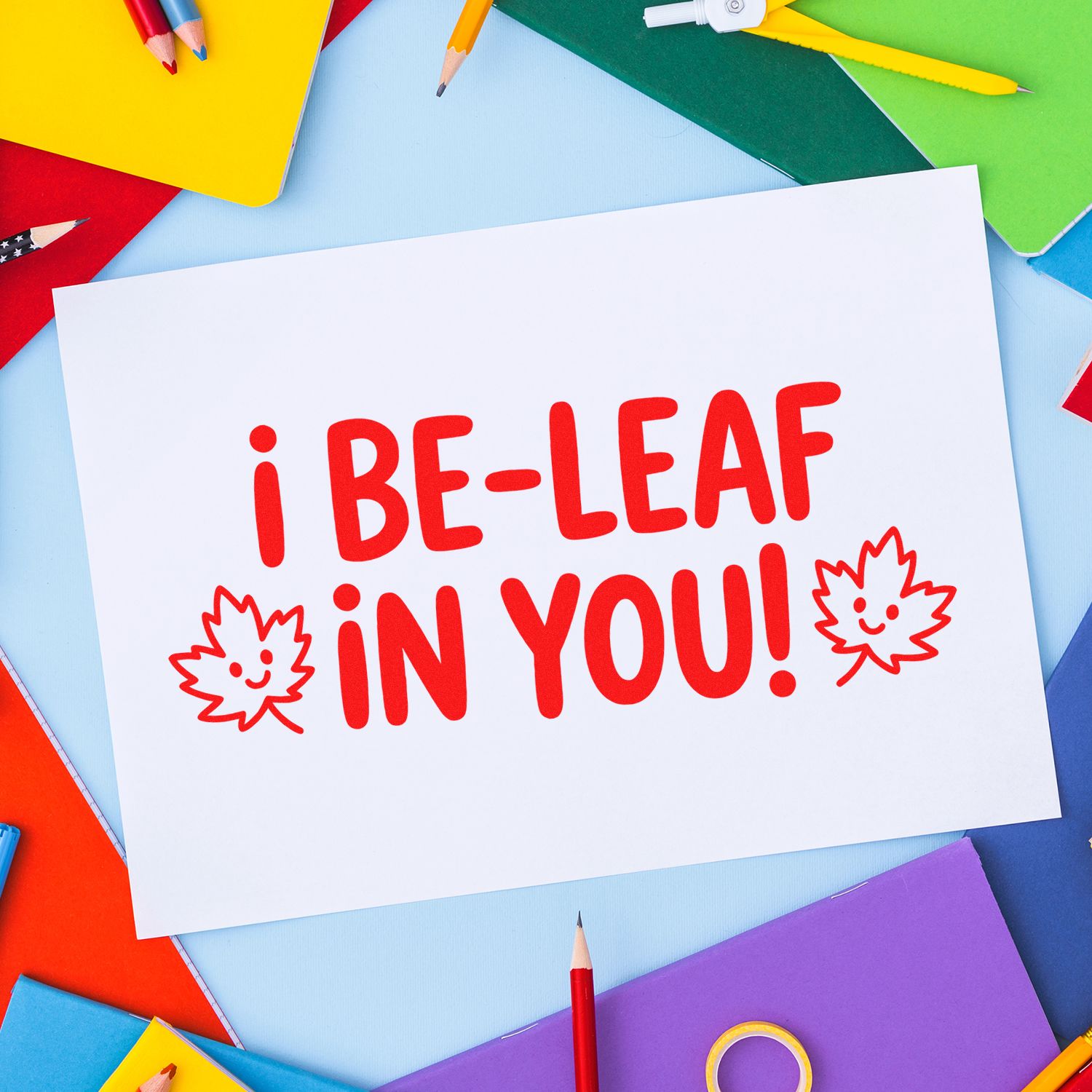 Red leaf stamp and seal message i BE-LEAF IN YOU! on white paper, with colorful pencils and notebooks.