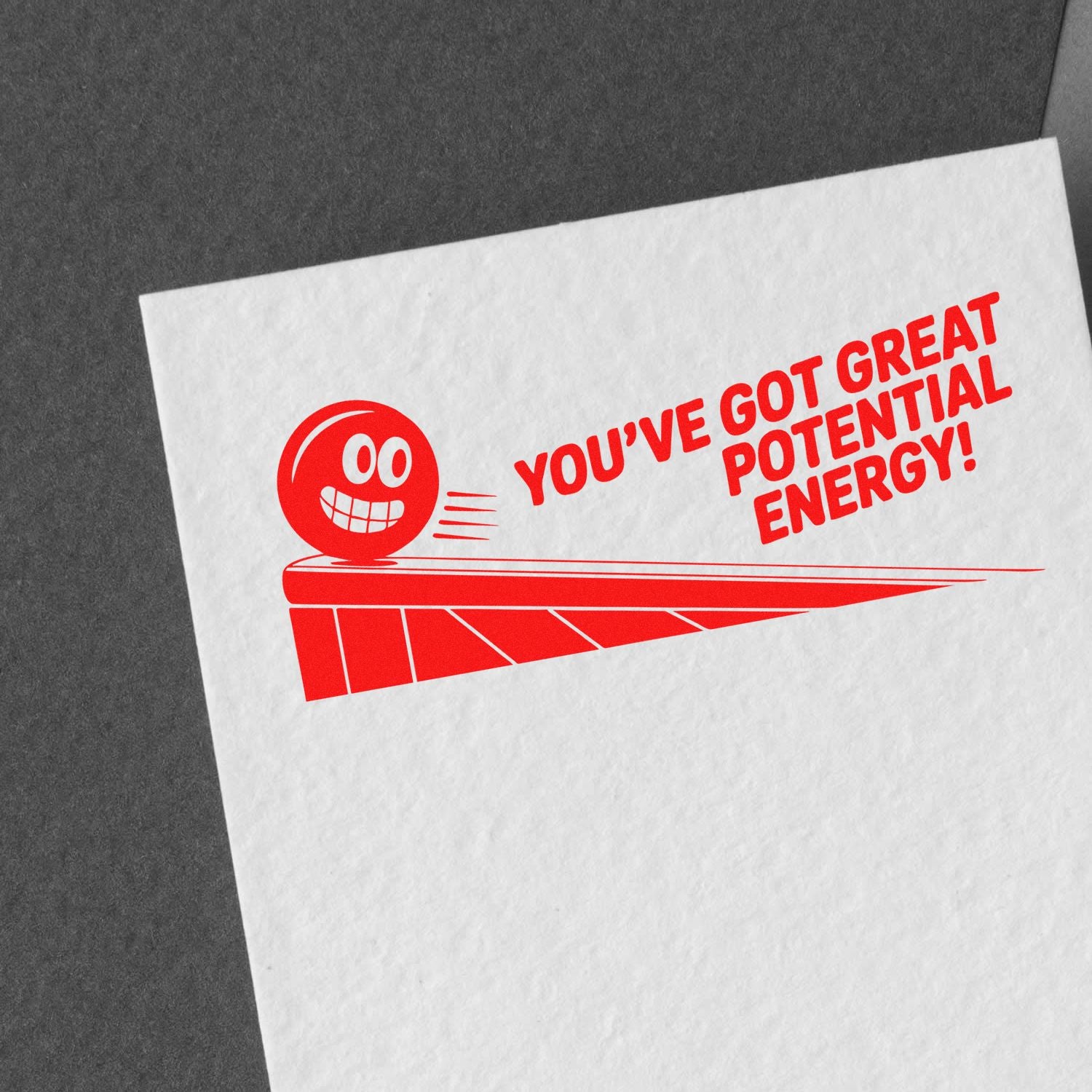Red rubber stamp seal with smiling face and Youve got great potential energy! on white paper, on a dark gray background.