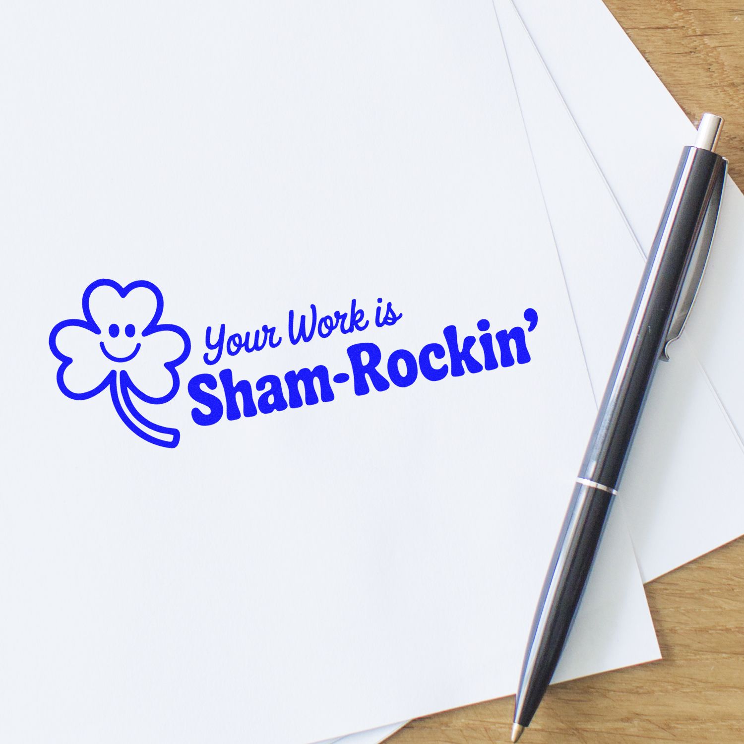 Blue clover-shaped rubber stamp reading Your Work is Sham-Rockin on white paper beside a black pen.