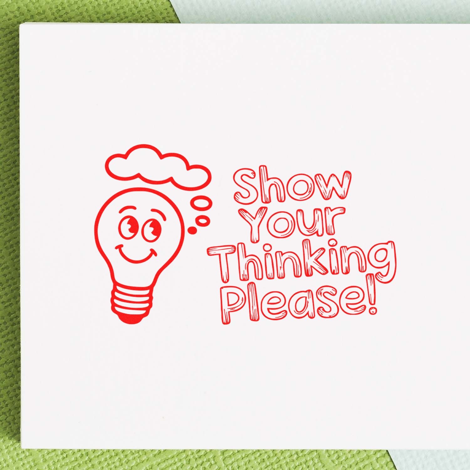 Red rubber stamp seal with smiling lightbulb and Show Your Thinking Please! on white paper over a green background.
