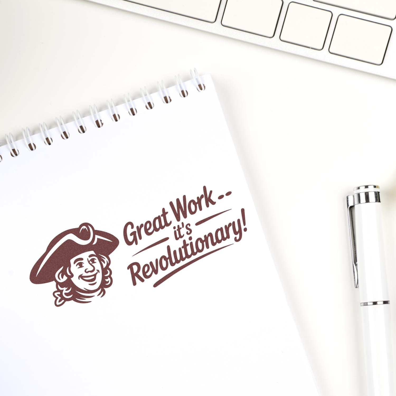 Brown rubber stamp and seal on white notebook reading Great Workits Revolutionary! beside a white pen and keyboard.