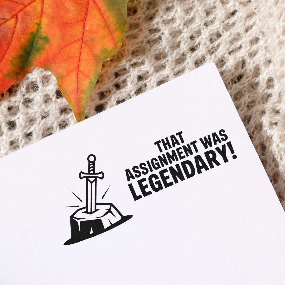 Black stamp seal on white card reads That assignment was legendary! with sword-and-stone icon, on a beige knit background with autumn leaf.