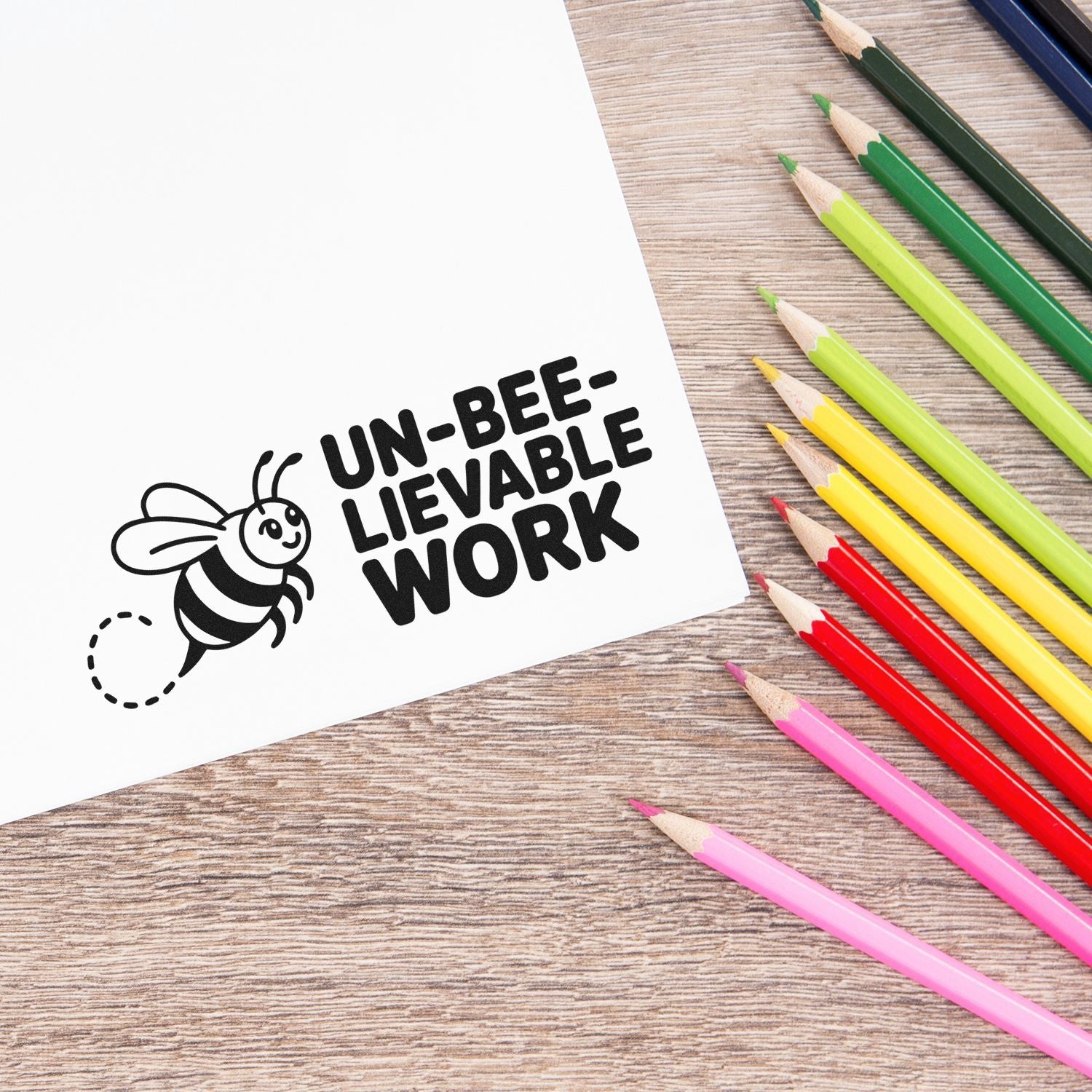 Black bee stamp seal with UN-BEE-LIEVABLE WORK on white paper, surrounded by colorful pencils on a wood desk.