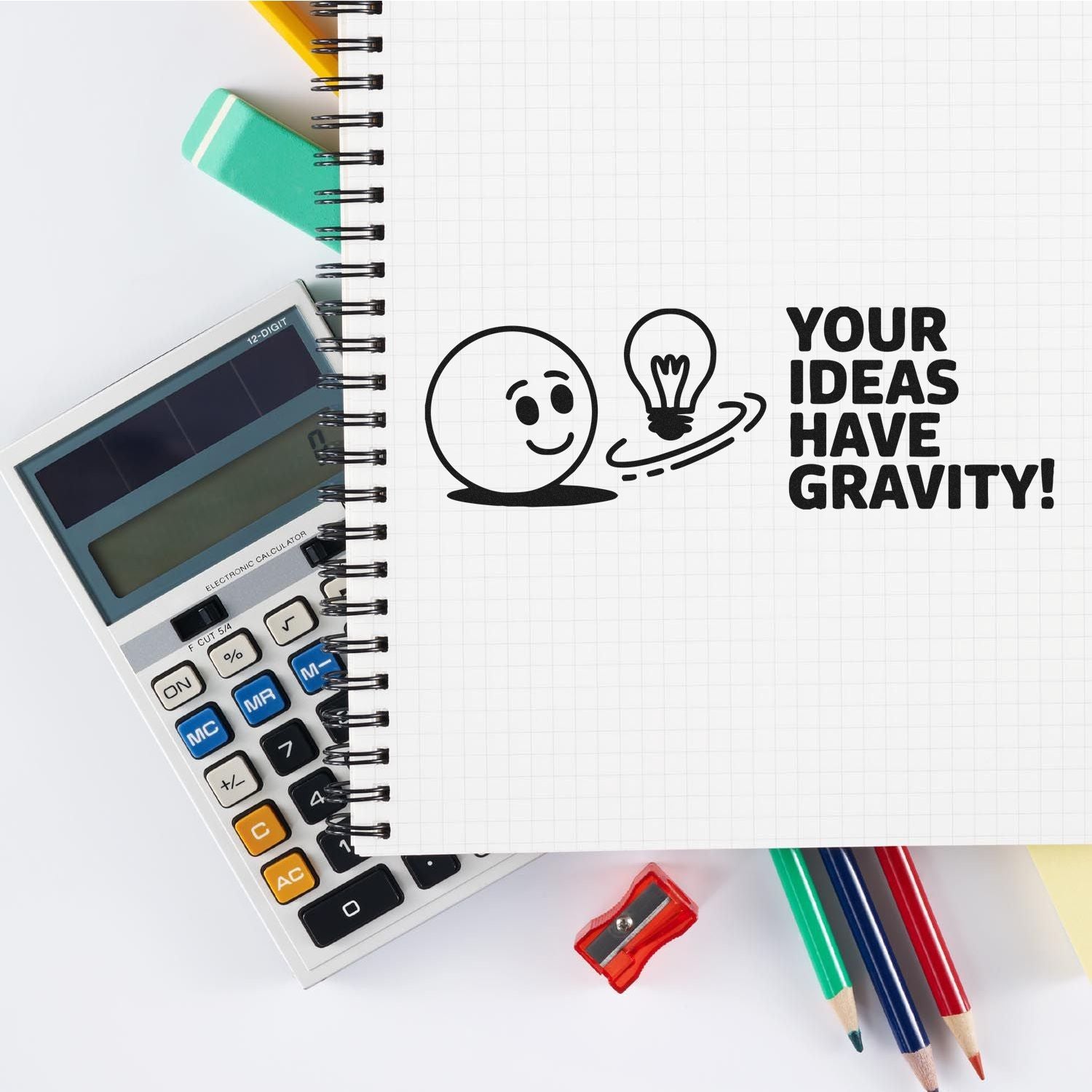 Black stamp seal on notebook reads Your Ideas Have Gravity! with lightbulb and smiley, beside calculator, pencils and sharpener.