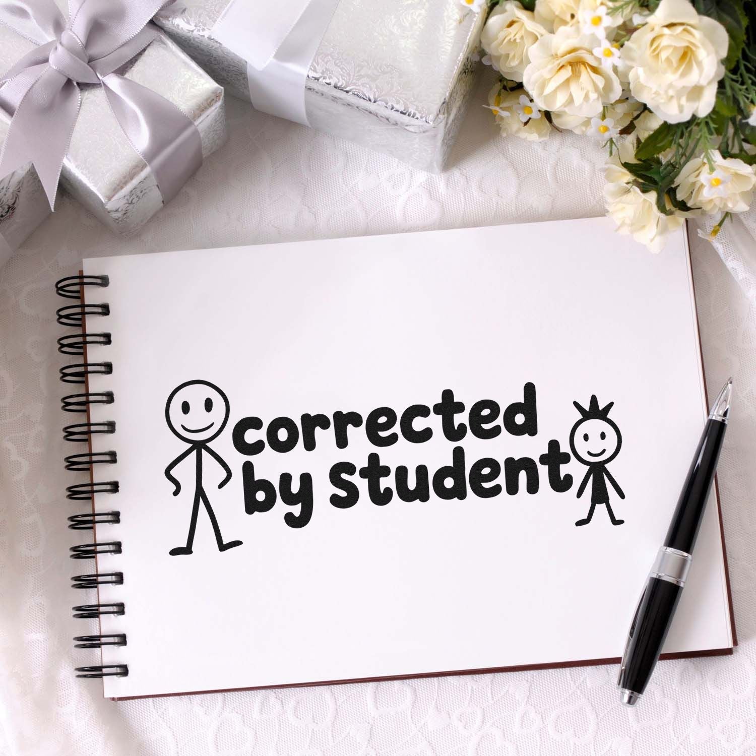 Black corrected by student stamp on a white spiral notebook, with pen, silver gift boxes, and cream flowers.