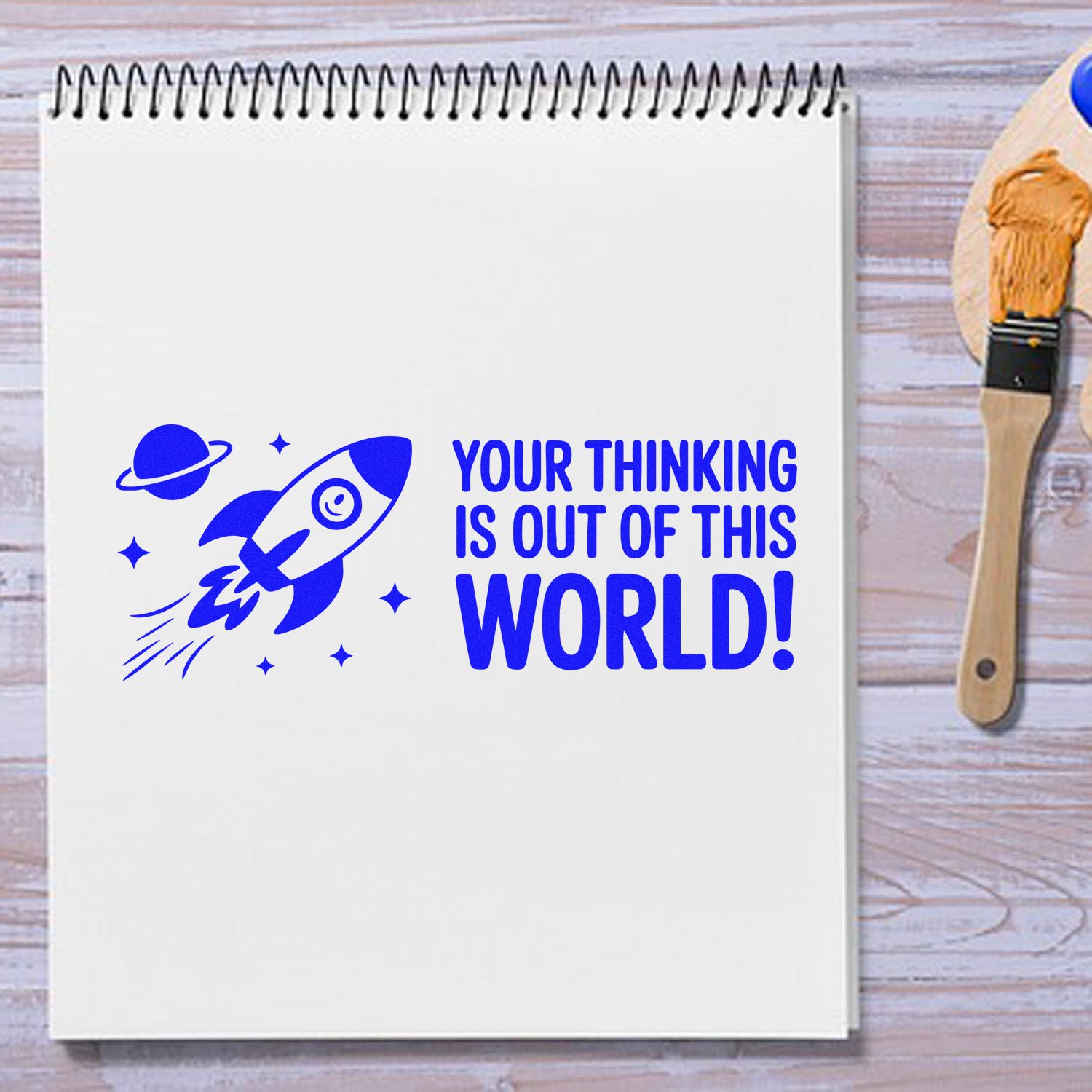 Blue rubber stamp and seal design with rocket and Your thinking is out of this world! on a spiral notebook, blue ink, paintbrush.
