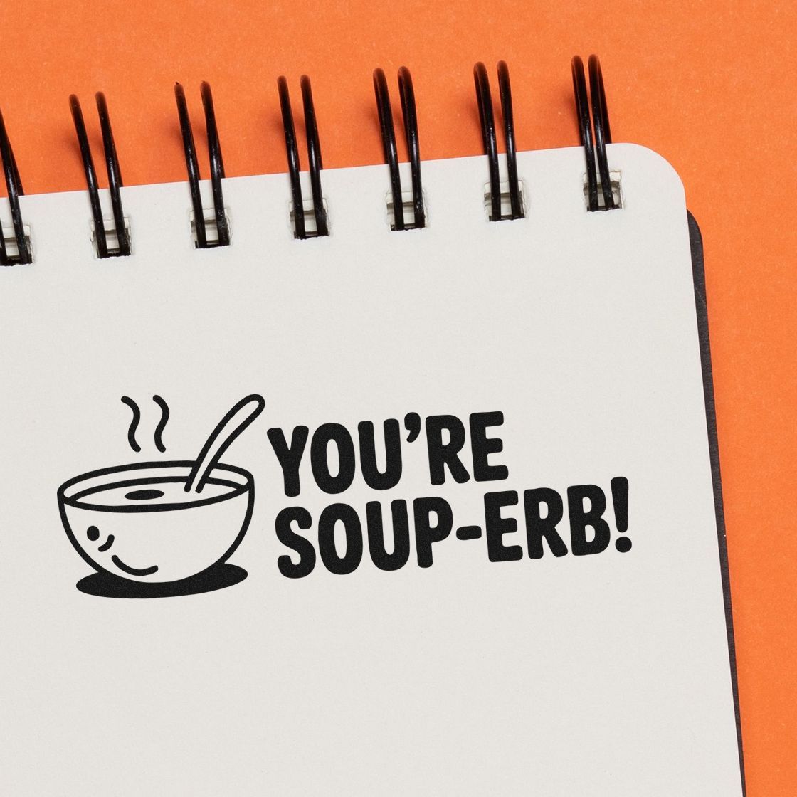 Black soup-erb stamp and seal with smiling soup bowl icon on white notebook, against a bright orange background.