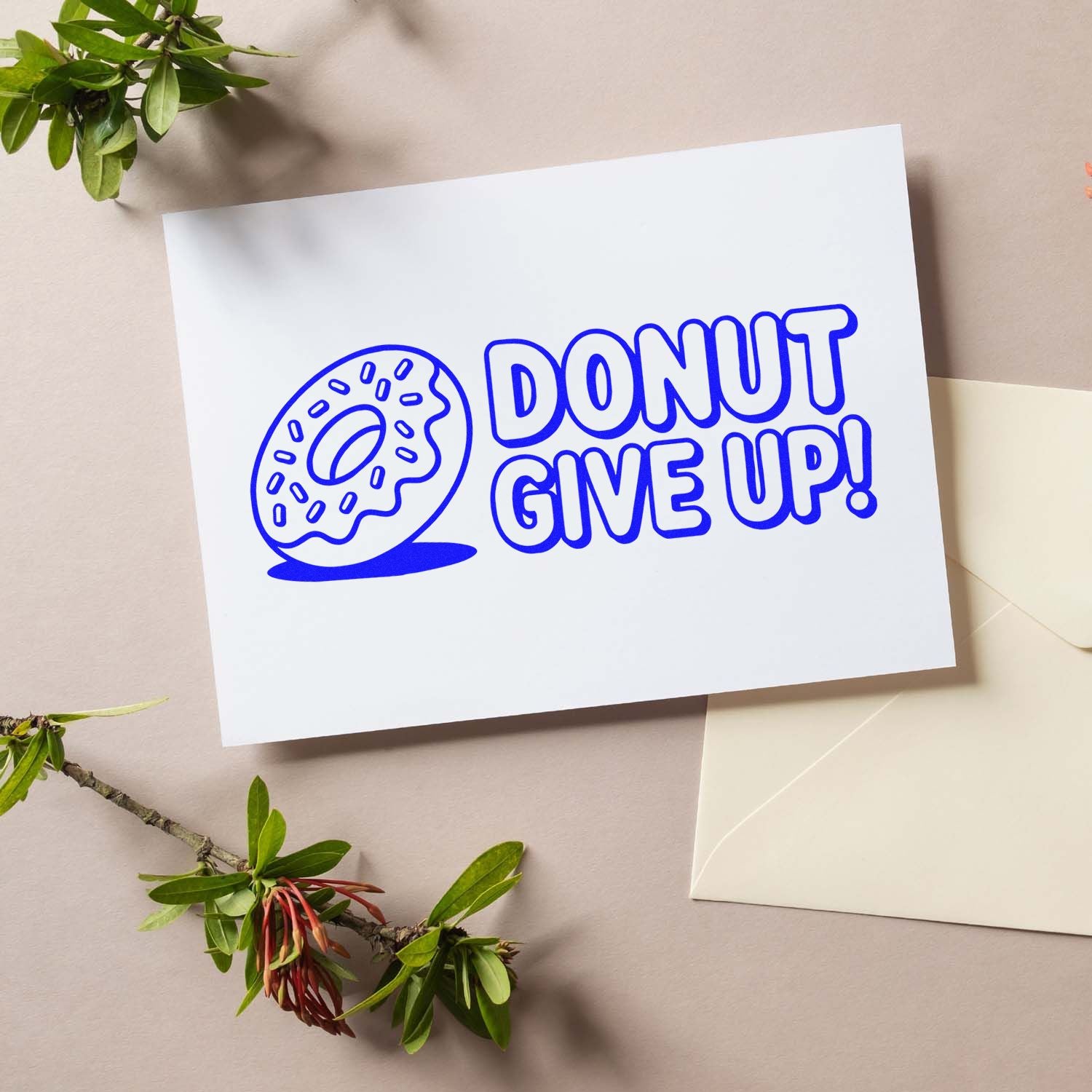 Blue donut stamp-style seal reading DONUT GIVE UP! on white card, with cream envelope and green leaves.