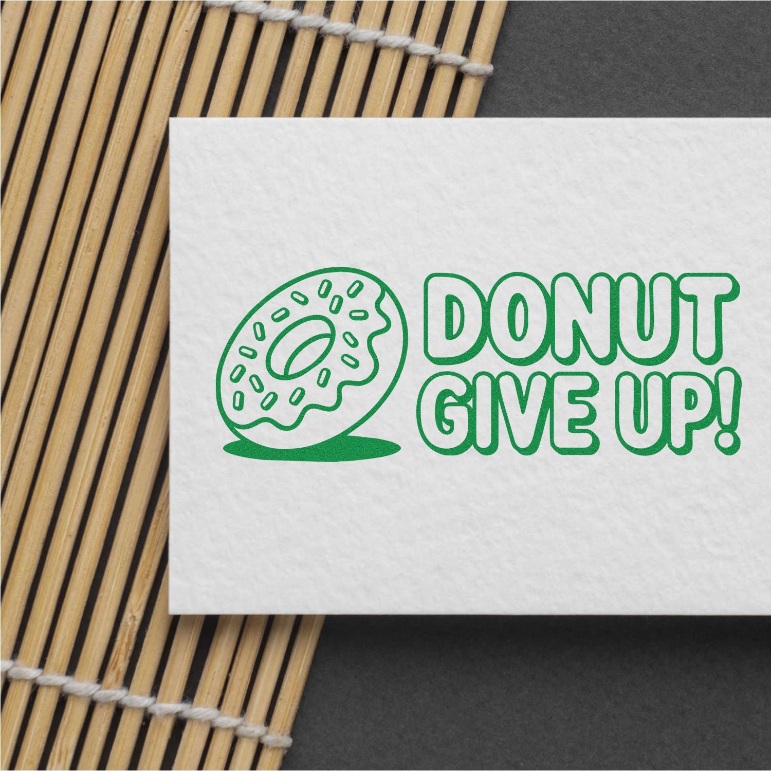 Green donut stamp and seal design reading DONUT GIVE UP! on white paper over a bamboo mat.