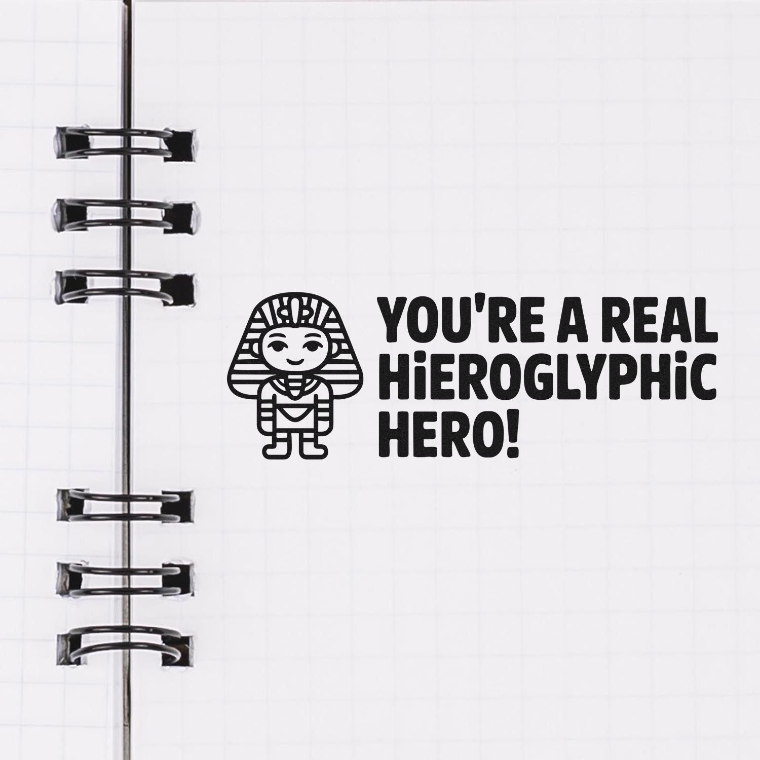 Black hieroglyphic hero stamp and seal with pharaoh icon and bold text on white notebook page, beside silver binder rings.