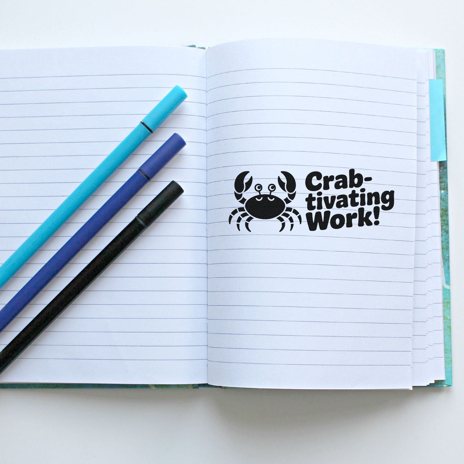 Black crab stamp/seal reading Crab-tivating Work! on lined notebook with teal, blue, and black pens.